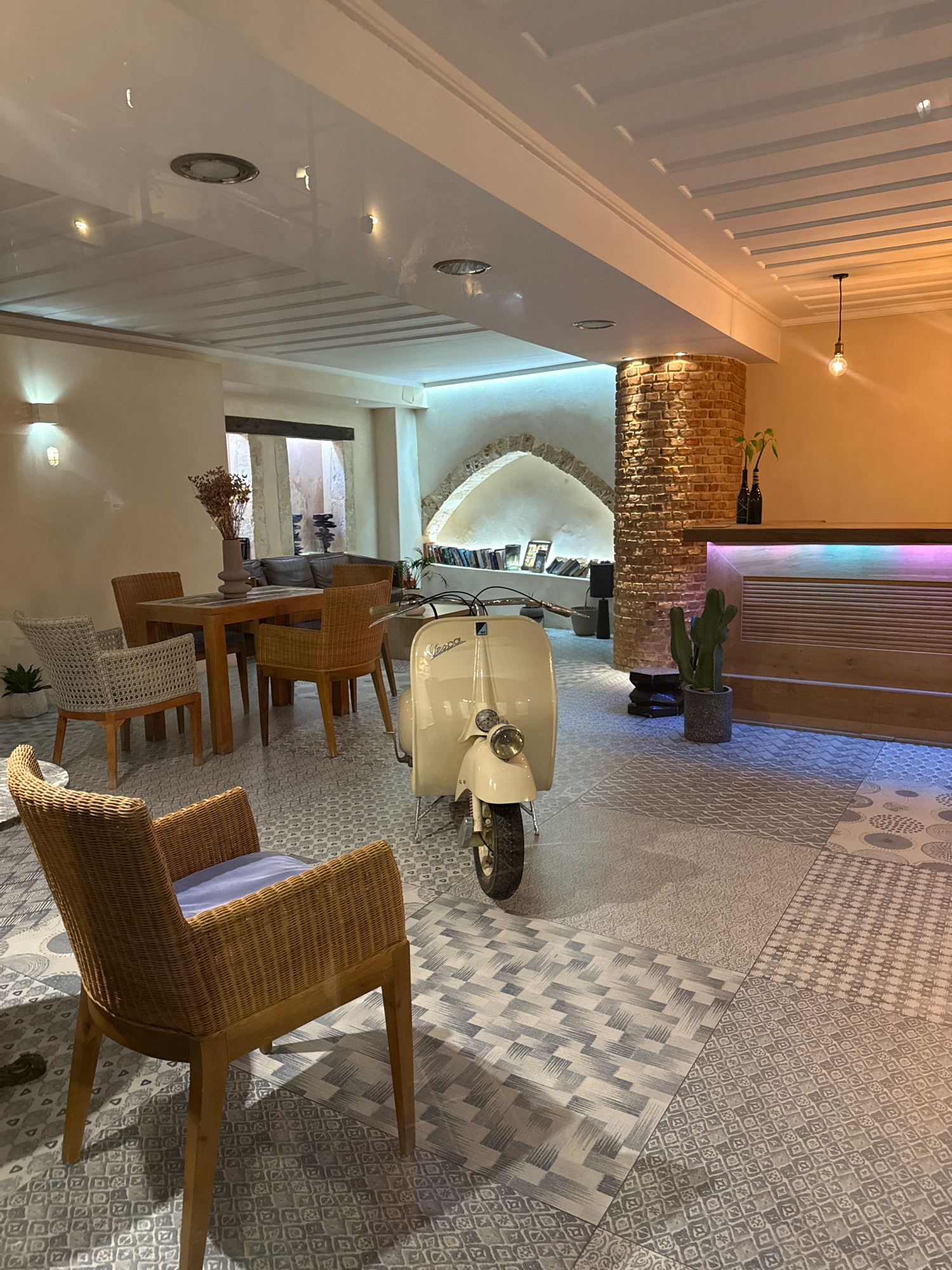 The image depicts the interior of a well-lit establishment, possibly a cafe or hotel lobby, located in an unknown city and district. In the foreground, a cream-colored Vespa scooter is parked on a floor adorned with a patchwork of patterned tiles in various shades of grey and white. To the left, a wooden dining table is surrounded by four chairs: two made of woven wicker with light-colored cushions, and two constructed from woven rattan. A vase holding dried flowers sits on the table. Further back, a reception desk with a wooden front and a multicolored illuminated top is visible, with a potted cactus placed to its left. The background reveals a brick-clad pillar and an arched entryway leading to a space with bookshelves. The ceiling features a modern design with recessed lighting and exposed beams, and a single pendant light hangs from the right side. The walls are painted in neutral tones, with one section of exposed brick and another with a textured, light-colored finish.