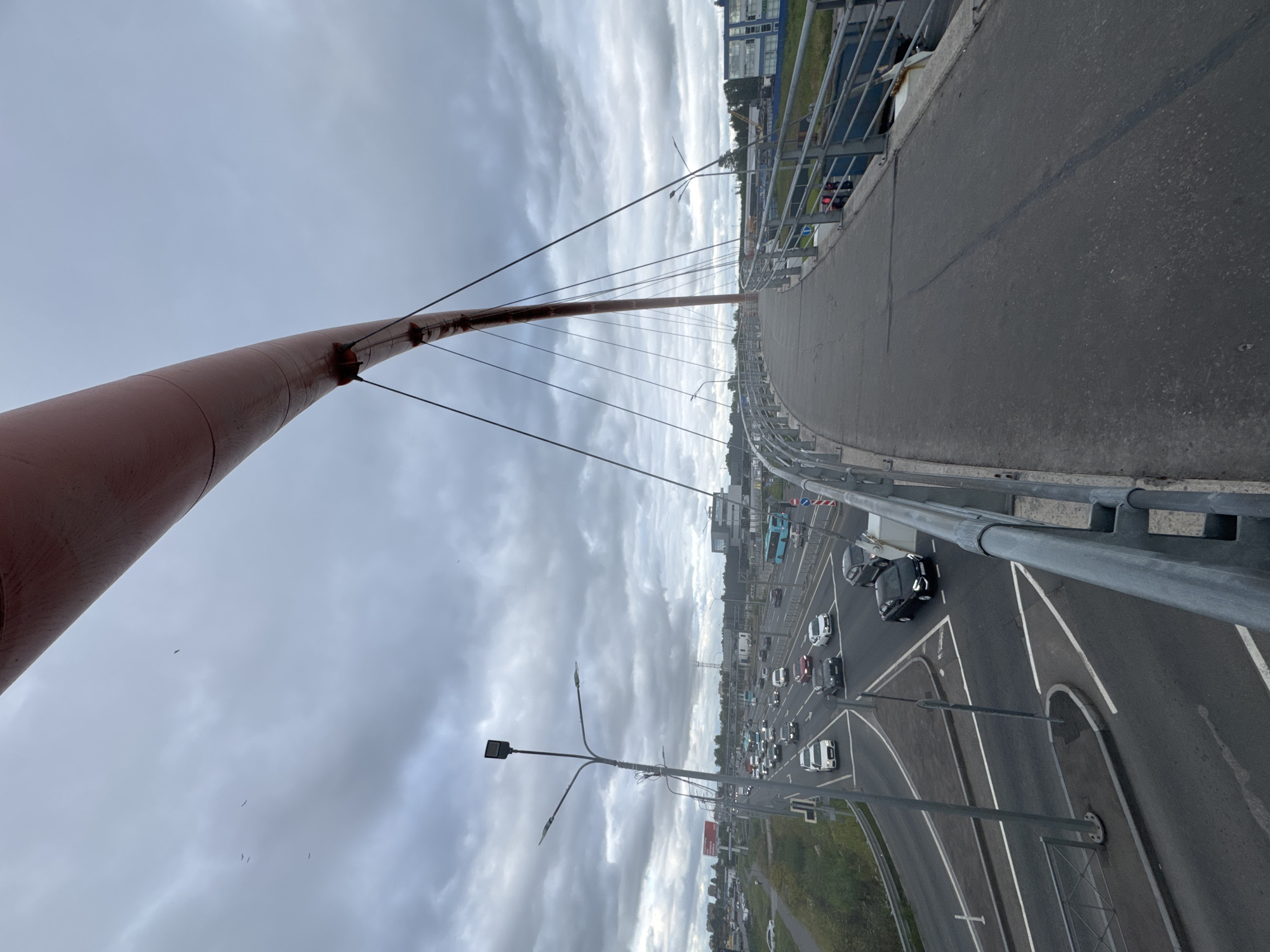 The image depicts a slightly overcast day from the perspective of a pedestrian walkway on a modern bridge in Saint Petersburg, Russia. The dominant subjects are the bridge structure itself, characterized by its curved asphalt surface, metal railings, and a distinctive red pylon supporting suspension cables. Below the walkway, a multi-lane road carries a moderate amount of traffic, including cars and a blue bus. The scene suggests a typical day with urban activity.
Notable details include the cloudy sky, which hints at a potentially cool or damp weather condition, common in this region. The time of day appears to be daytime, given the visibility of the road and traffic. The architecture of buildings in the background suggests an urban environment, likely an industrial or commercial area given the presence of larger structures and the highway. The image is shot with a wide-angle lens, giving a sense of scale and depth. No discernible text is visible in the image. The overall mood is neutral, focusing on the infrastructure and the flow of traffic under a diffused sky. The presence of some birds in flight against the sky adds a touch of natural element to the urban landscape.
