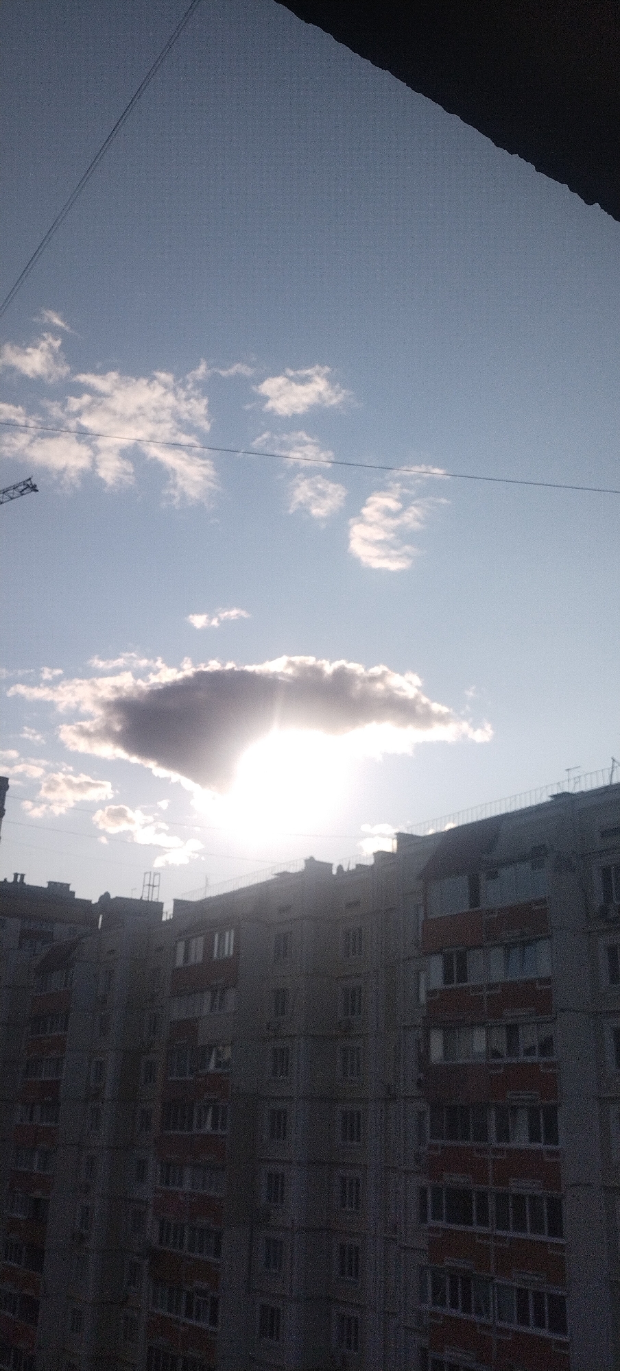 The image shows the sky above a residential building in Pisochyn, Ukraine, possibly during sunset or sunrise due to the sun being low in the sky and partially obscured by a cloud. The sky is a pale blue, with scattered white and grey clouds. A thin dark line, likely a power line or wire, runs diagonally across the upper portion of the image. Below the sky, a multi-story building with numerous windows and balconies dominates the lower half of the frame. The building appears to be made of concrete and has a gray facade, with some sections of balconies painted red. The overall scene is urban and residential.