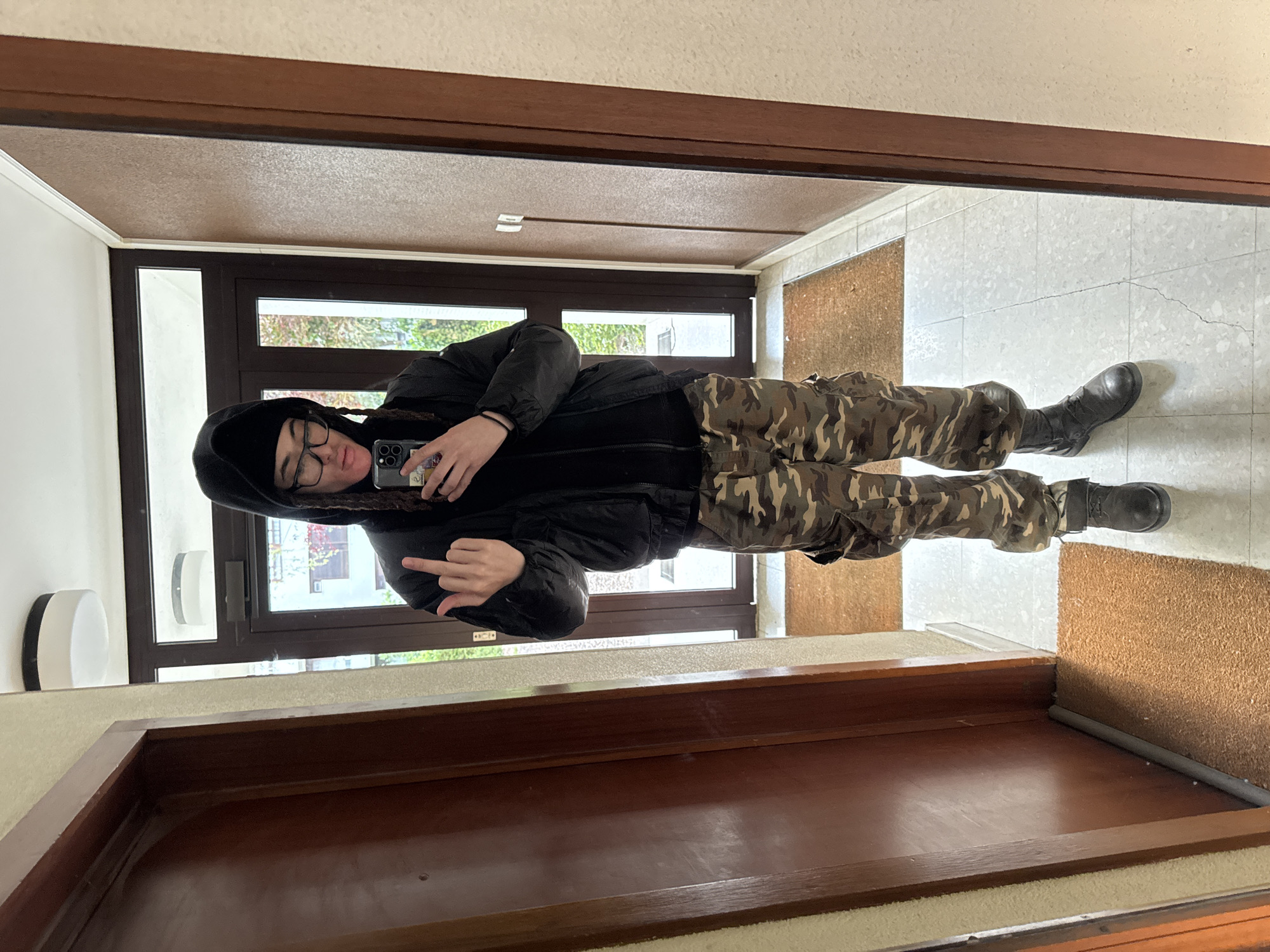 An individual is captured in a mirror selfie within what appears to be an indoor entryway or hallway in Les Ulis, France. The person, appearing young, is seen from the chest down, wearing a black beanie, clear-framed glasses, a black hooded jacket, camouflage-patterned pants, and black combat boots. Braids are visible on the side of their head.
They are holding a smartphone (possibly an iPhone) in their right hand to take the photo, and their left hand is raised, making a middle finger gesture towards the camera. The expression on their face, partially obscured by glasses and the pose, seems neutral, contributing to a defiant or playful demeanor.
The setting, reflected in the mirror, features a light grey tiled floor with visible cracks, a textured brown doormat, and a large wooden door on the right. Behind the person, a wide window with dark frames provides a glimpse of an outdoor scene with green foliage and distant buildings, indicating it is daytime. A white, circular light fixture is mounted on the wall to the left of the subject. No legible text is discernible in the image.