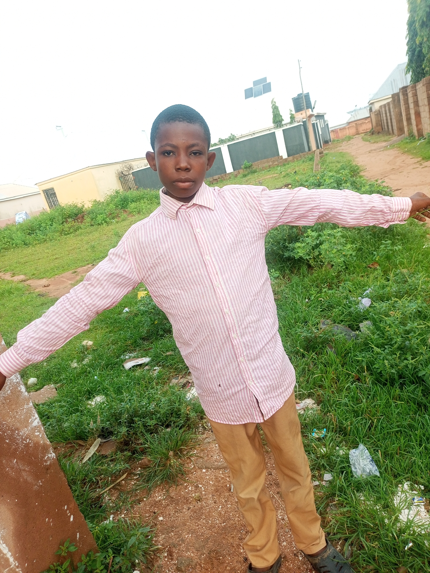 A young boy stands outside, with his arms outstretched. He is wearing a pink and white striped long-sleeved button-up shirt and tan pants. The background shows a dirt path, grass, some buildings, and a brick wall. The sky is overcast. The location is Tempure, Nigeria.