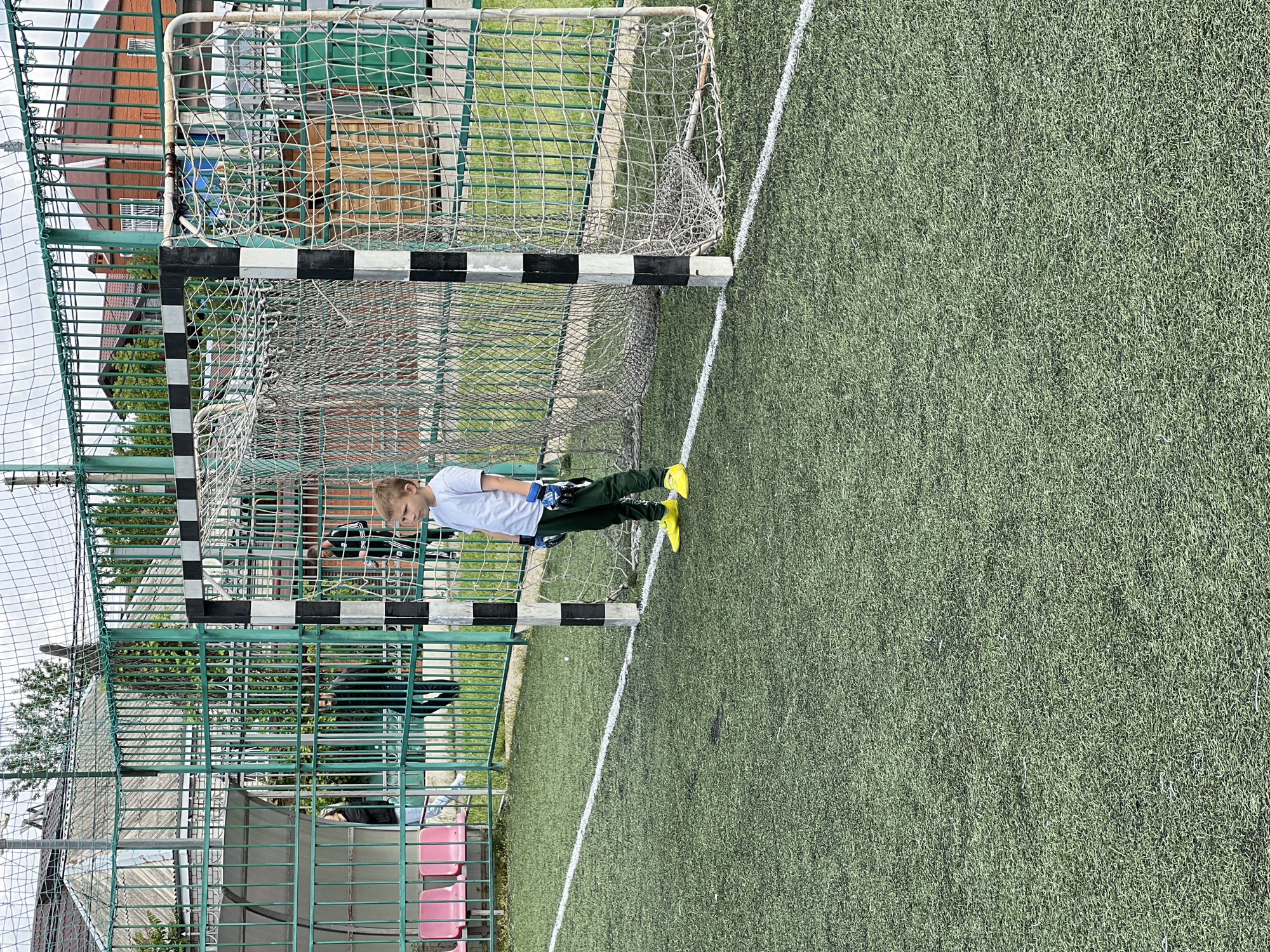 A young person in goalkeeper gear stands on a green athletic field next to a soccer goalpost with black and white stripes, likely in Krasnodar, Russia, given the architectural styles and structures visible in the background. The goalpost sits along a white line on the artificial turf. A metal fence borders the field. A section of seating, with pink seats, is also visible near the fence. In the background, residential buildings and trees can be seen. The sky is overcast.