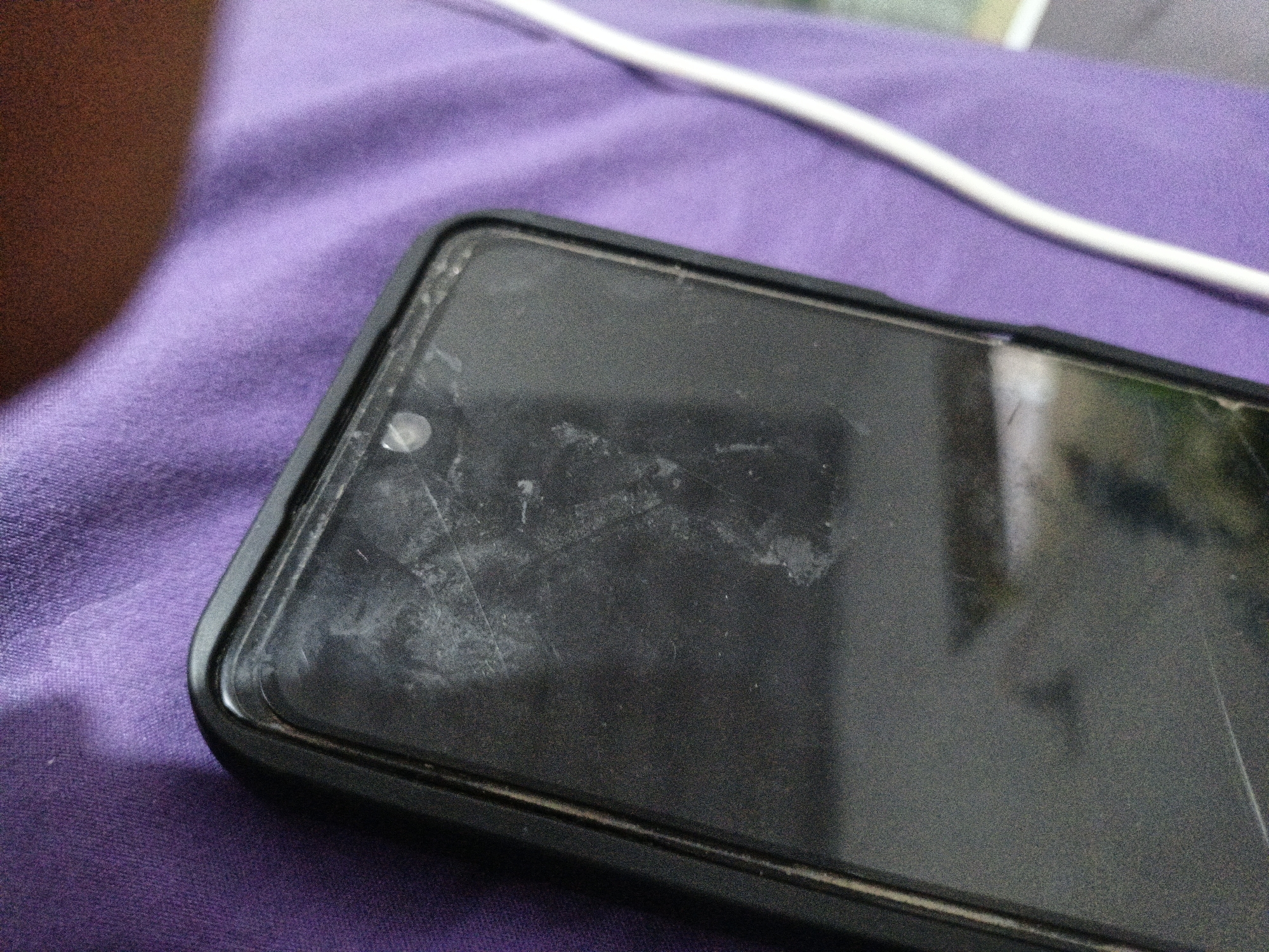 A close-up, slightly angled shot shows a smartphone with a black case lying on a purple, textured surface. The phone's screen is visible, appearing slightly smudged with white residue and faint scratches. A thin white cable runs diagonally across the top right of the frame, resting on the purple surface. The environment is indistinct beyond the immediate focus on the phone and the purple material. There are no people present in the image, and no discernable location within Teumpokteungku, Indonesia.