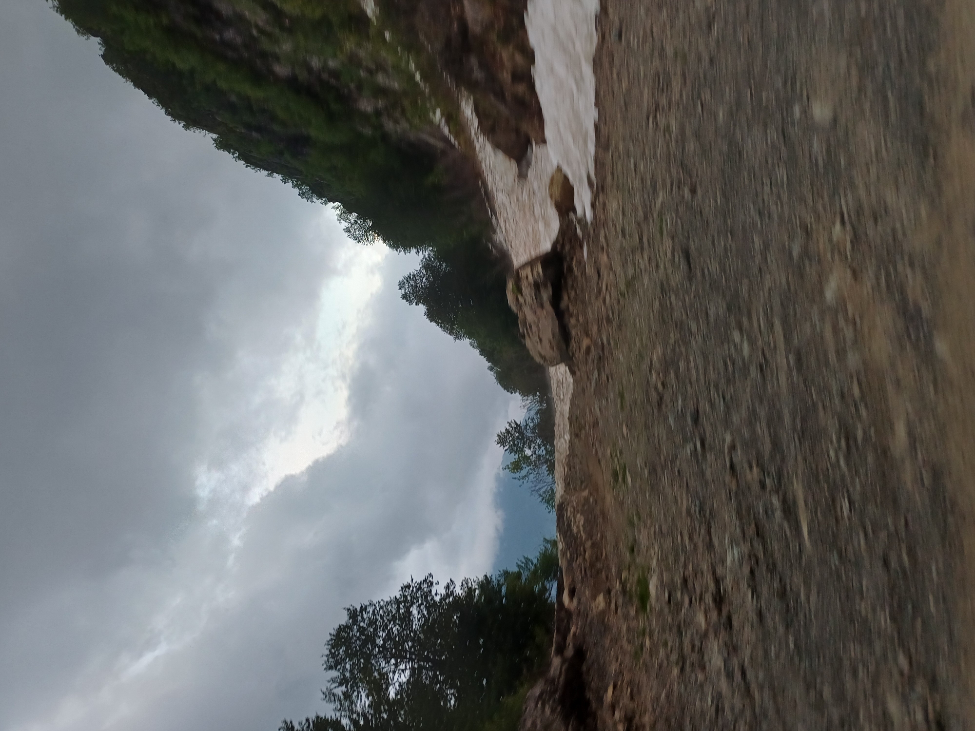 The image depicts a mountain road in Estosadok, Russia, under a cloudy sky. The foreground is a rough, gravelly road. To the left, a dense forest of dark green trees rises up a rocky incline. To the right, a steep, heavily forested mountainside dominates the scene, with patches of snow visible at its base. A large boulder rests on the snow-covered ground near the base of the mountain. The overall atmosphere is somber and overcast.