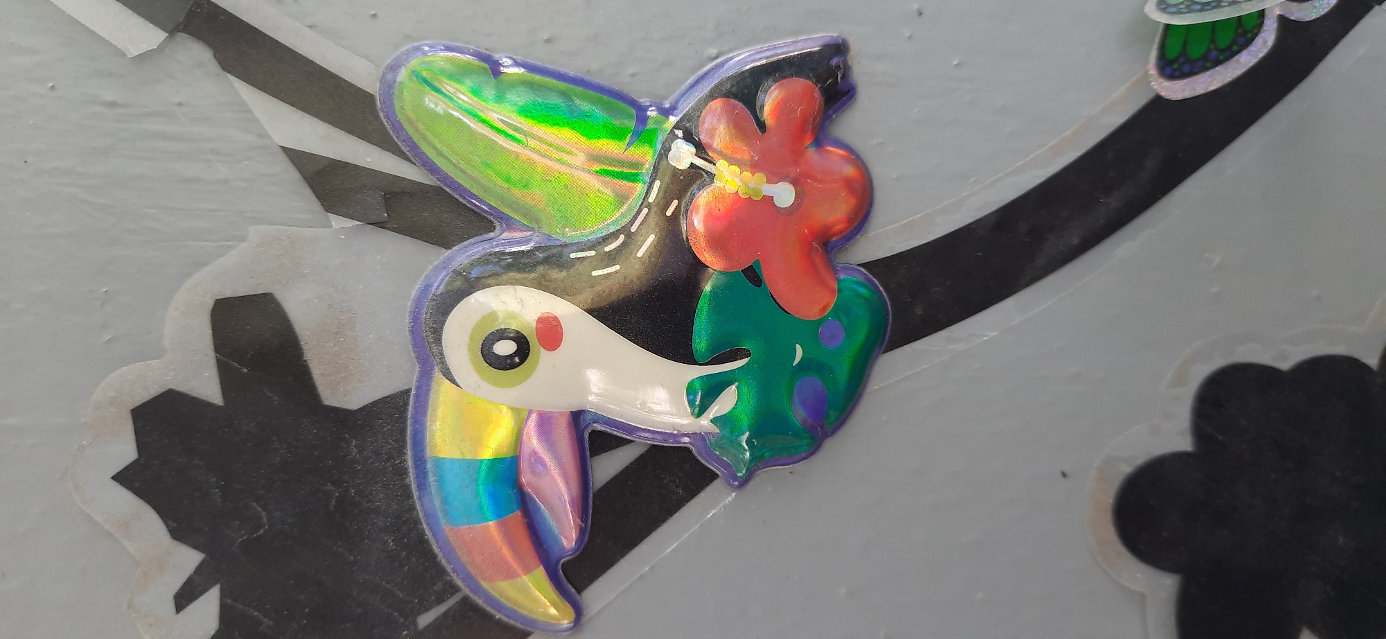 A holographic sticker depicting a cartoon toucan is affixed to a light gray, textured surface. The sticker features a white toucan head with a large, rainbow-colored beak. The toucan has large, green eyes with black pupils and rosy red cheeks. It has a black body with iridescent green wings. Below the toucan's head, there are green, leaf-like shapes with purple polka dots. A red flower with a yellow center is positioned to the right of the toucan. The background behind the sticker consists of irregular black and white striped shapes. The sticker appears to be peeling in some areas, revealing a white border. This sticker is located on a wall or structure in Dnipro, Ukraine.