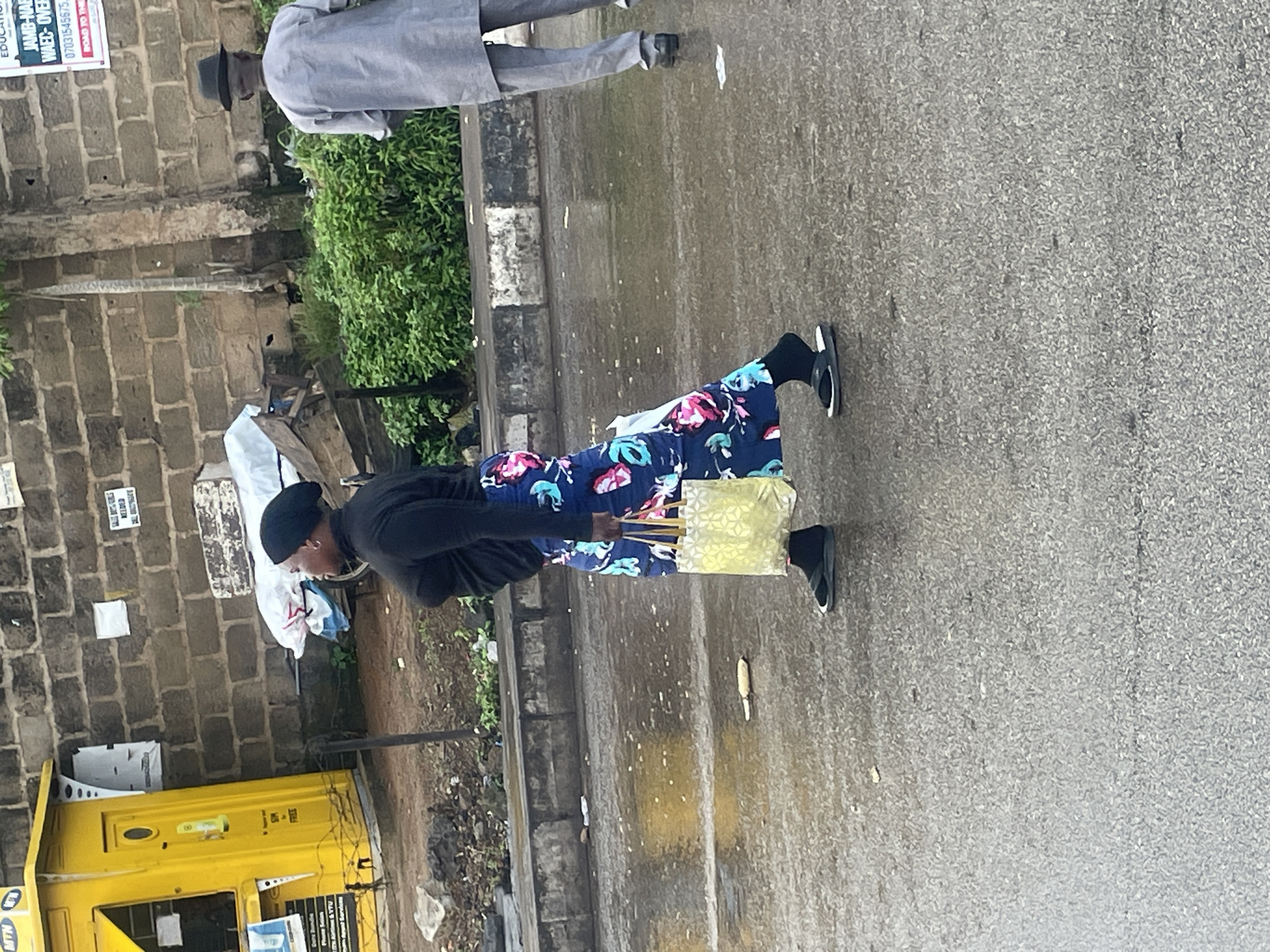 In Barakin Lerawi, Nigeria, a woman walks on a wet asphalt road carrying a patterned tote bag. She is wearing a black headscarf, a black turtleneck top, and a long, dark blue skirt with a floral pattern. She has on black socks and black flip-flops. To her right, a man in a grey kaftan and a black hat walks away from the camera. In the background, a yellow kiosk with the MTN logo stands next to a brick wall. The ground is wet and reflective, indicating recent rain. There is also some greenery and debris visible along the roadside.