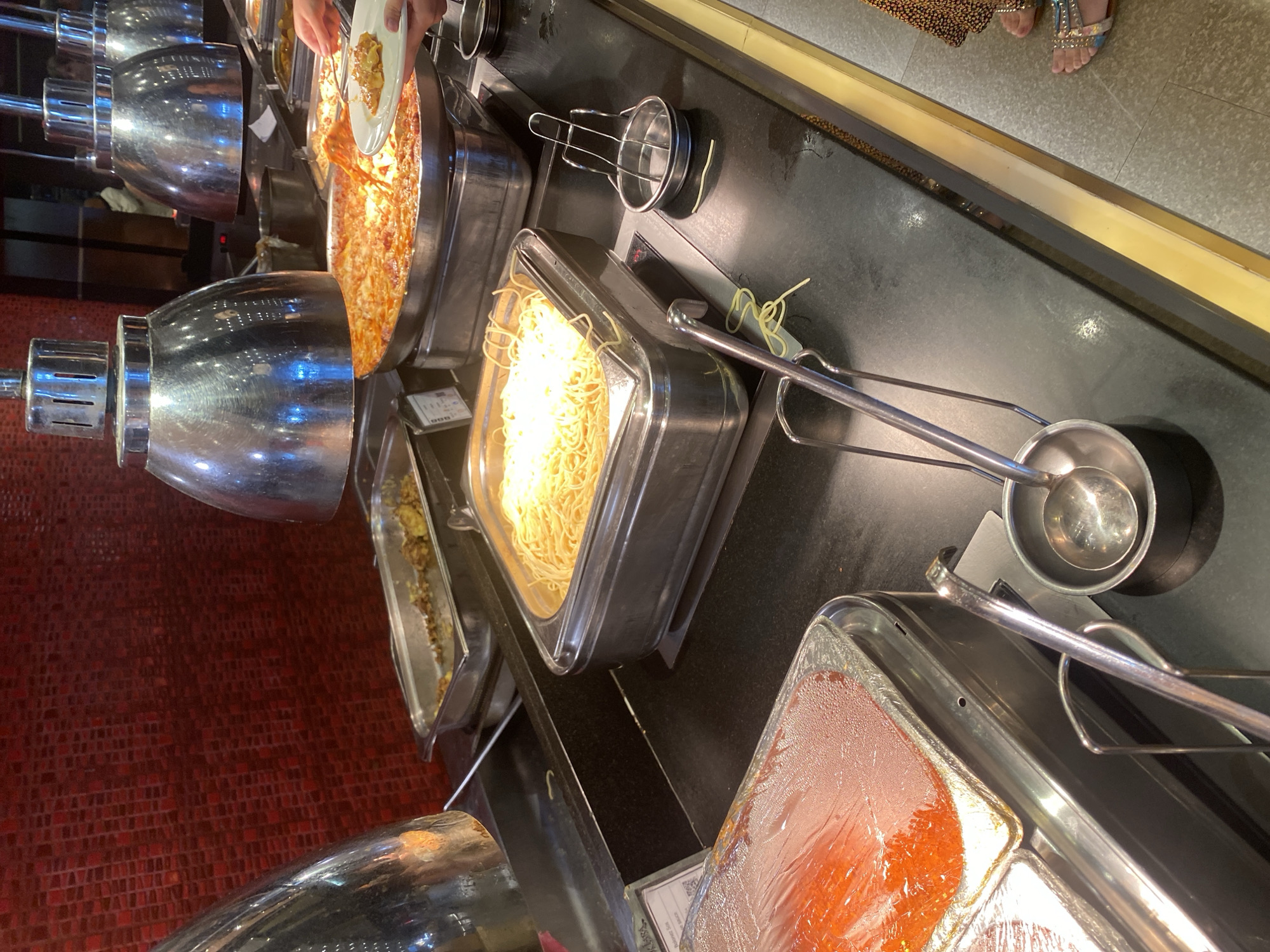 The image shows a buffet serving line, likely in a restaurant or hotel. In the foreground, there are two large stainless steel serving trays. The tray on the left contains a red sauce, possibly marinara or tomato sauce, covered with plastic wrap. The tray on the right holds a large portion of spaghetti. Next to the spaghetti tray, there are serving utensils, including a ladle in a small metal holder and a pair of tongs resting on a metal rack. In the background, a large pan of lasagna is visible, with a person's hand reaching in with a plate to serve themselves. Other serving dishes with food are present further back. The serving area is illuminated by overhead heat lamps. The background features a red tiled wall. There is no discernible information to place this scene in Aksu, Turkey.