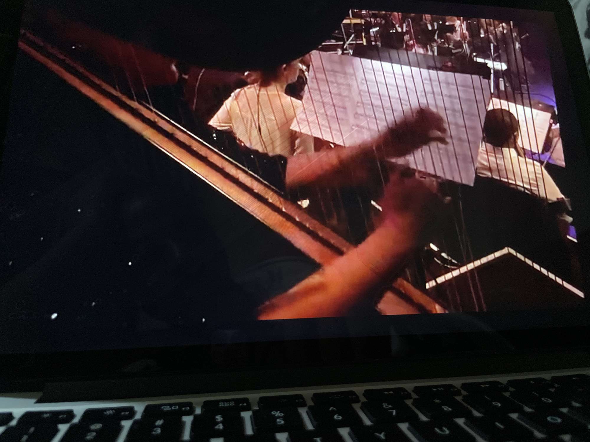 The image is a close-up, low-angle shot of a laptop screen displaying a video of a harpist playing in what appears to be a concert hall. The harp has a dark wood frame and a multitude of strings that reflect light. The harpist, wearing a light-colored shirt, is partially visible, with their hands in motion as they play. Behind the harpist, other musicians can be seen, suggesting an orchestra or ensemble performance. A large sheet of music is placed on a stand in front of the harpist. The foreground of the image shows the keyboard of the laptop, with some keys illuminated. The lighting in the video is dim, with spotlights illuminating the performers and the harp. The image does not contain any information that would allow for the identification of a specific location in Howayi, Nigeria.