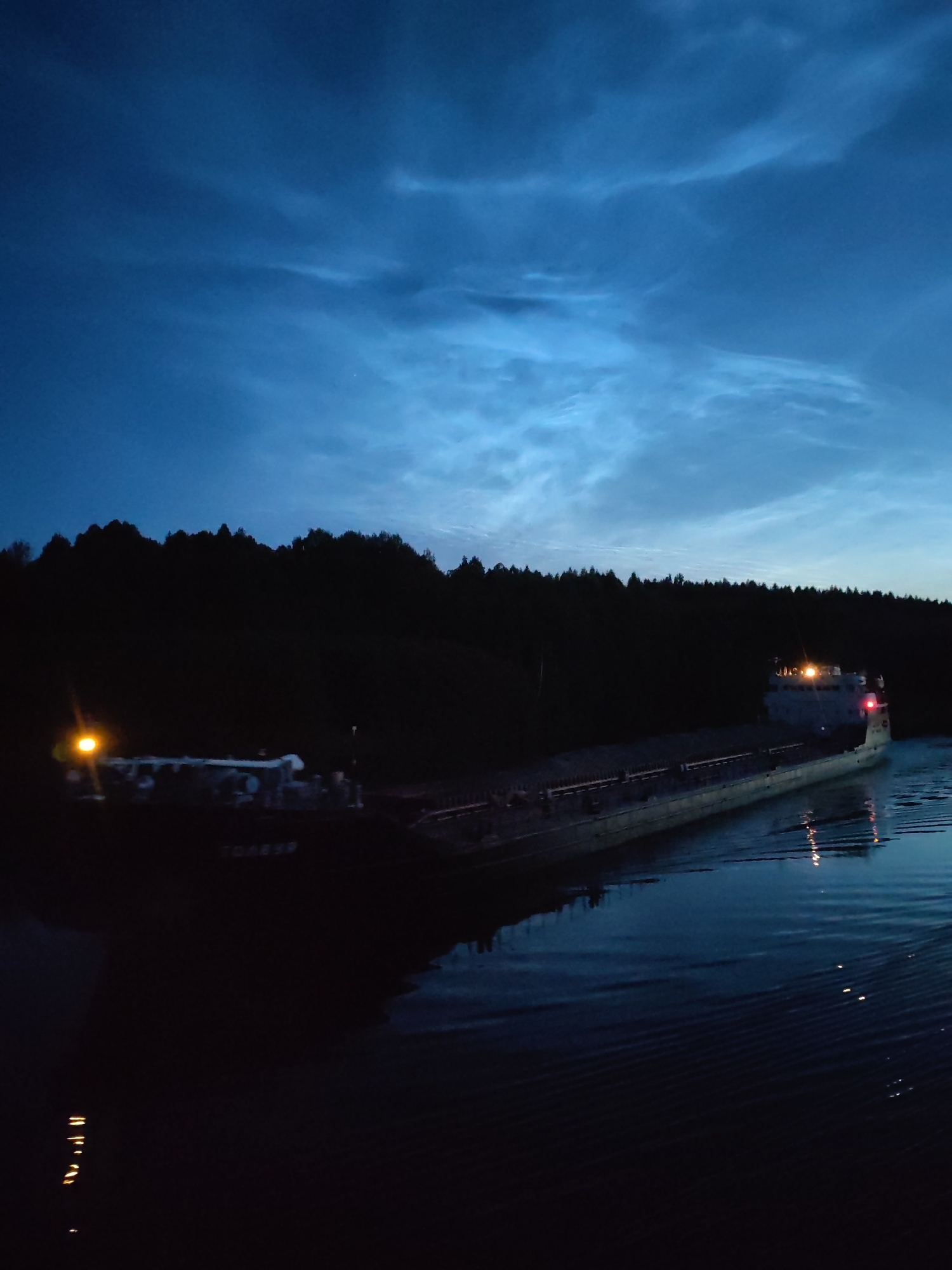 A cargo ship is navigating a body of water at dusk. The sky is a deep blue with wispy clouds. The ship is dark, with lights visible on its upper deck and at its stern. The ship appears to be moving towards the right side of the frame. Along the left bank of the water, there is a dark, silhouetted line of trees. The water is calm, with small ripples and reflections of the ship's lights. The scene is likely located in or near Volokov Most, Russia, a settlement on the Volkhov River.