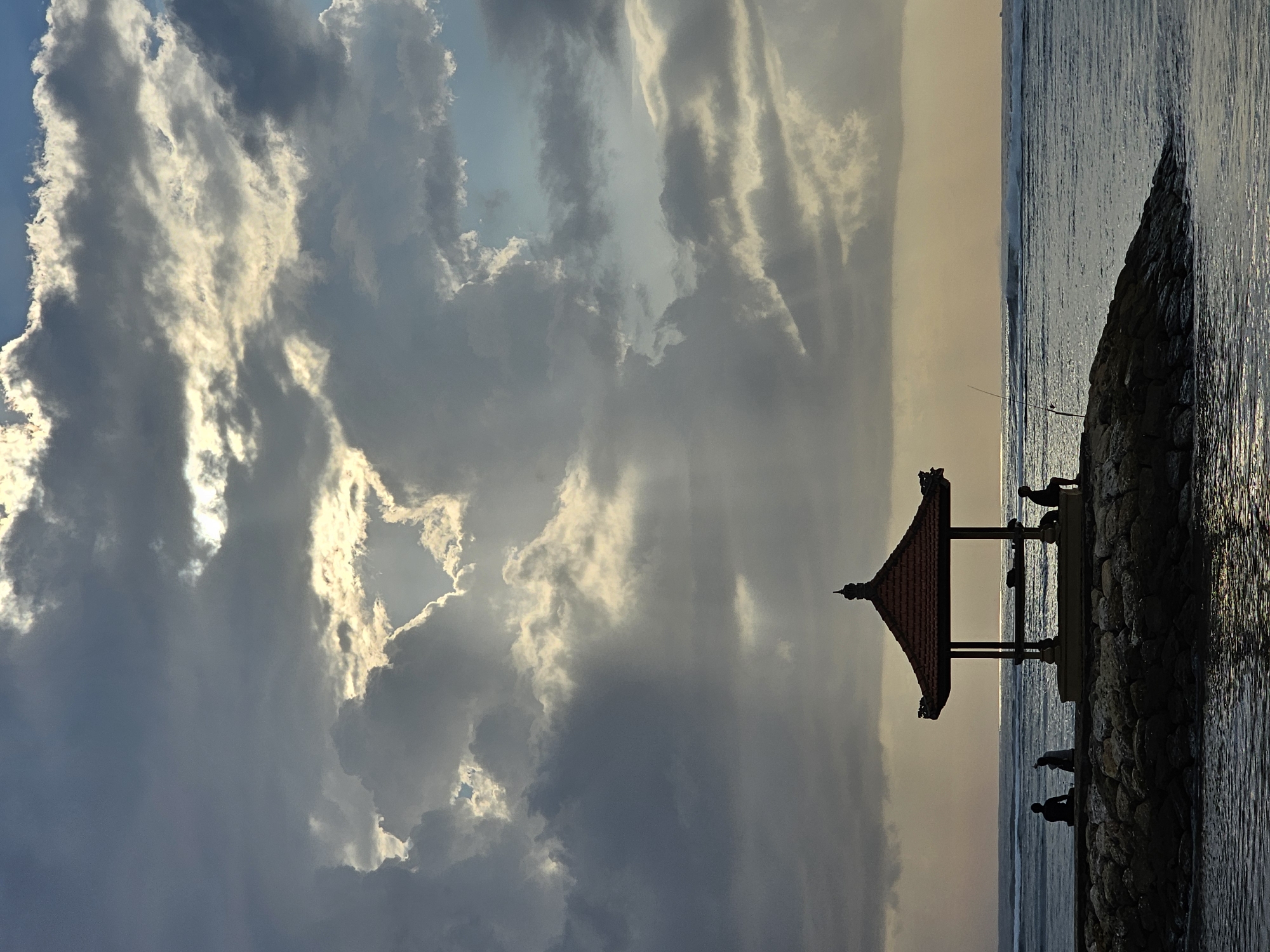 This image captures a serene scene on the coast of Denpasar, Indonesia, likely at Sanur Beach, during either sunrise or sunset. The sky is dramatic with heavy, gray clouds partially obscuring a bright, luminous sun, casting shafts of light down towards the ocean. In the foreground, a man-made stone jetty extends into the water, adorned with a traditional Balinese gazebo.
On the jetty, three figures are silhouetted against the bright horizon. To the left, two individuals sit close together, appearing contemplative. Further to the right, a third person is seated and appears to be fishing, with a fishing rod held upright. The gazebo, with its tiered roof and open structure, provides a focal point and a place of shelter. The ocean is calm, with gentle waves rolling towards the shore, and the water reflects the muted light of the sky. The overall atmosphere is one of peace and tranquility, a moment of quiet reflection by the sea.