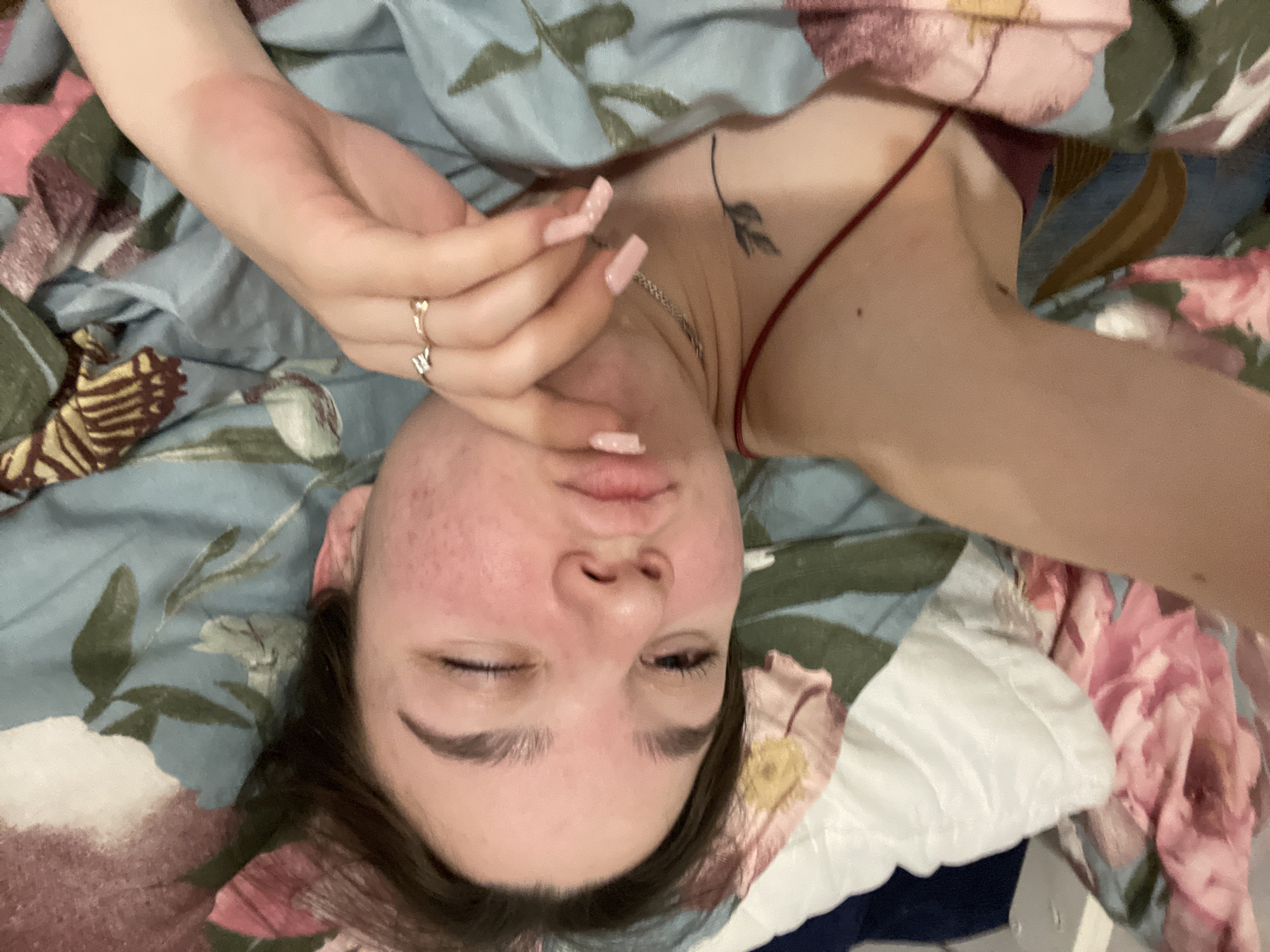 A young woman is lying on her back on a bed with a floral patterned duvet and pillow in an indoor environment. She is wearing a thin-strapped top. The woman has fair skin, dark eyebrows, and a small tattoo of a leaf or flower on her chest near her neck. She wears several rings. Her face has some redness and possible acne. Her hand is raised near her face. The exact location is unknown but hypothesized to be Ольховка, Russia.