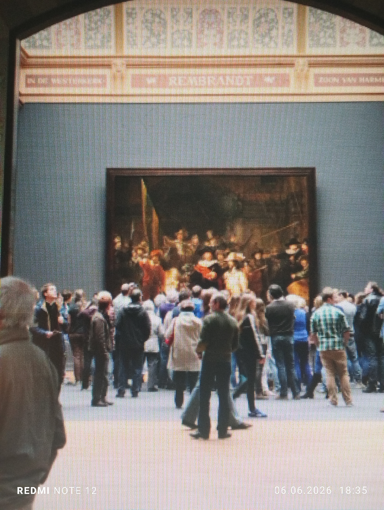 The image displays an interior view of an art museum gallery, centered on a large oil painting, clearly identifiable as Rembrandt's "The Night Watch." The painting is housed in a dark frame against a plain, medium-grey wall. Above the painting, a decorative architectural element features a light-colored band with legible text, including "REMBRANDT" and partially obscured Dutch text like "ZOON VAN HARMS..." further confirming the subject and artist. Above this band, a patterned wall covering extends towards an arched ceiling structure at the top of the frame.
A substantial crowd of people is gathered before the painting, predominantly viewed from behind or in profile, indicating their focus on the artwork. The individuals wear varied casual clothing, including jackets, shirts, and trousers. A person in a light-colored top is partially visible in the immediate left foreground, facing away from the camera. The floor is a light, neutral color.
The image itself exhibits a prominent pixelated grid pattern across its entire surface, suggesting it is either a photograph of a digital screen or a low-resolution digital capture. A "REDMI NOTE 12" watermark is visible in the bottom left corner, and a date and time watermark "06.06.2026 18:35" is present in the bottom right corner.
Based on the distinct identification of "The Night Watch" by Rembrandt, the location is the Rijksmuseum in Amsterdam, Netherlands.