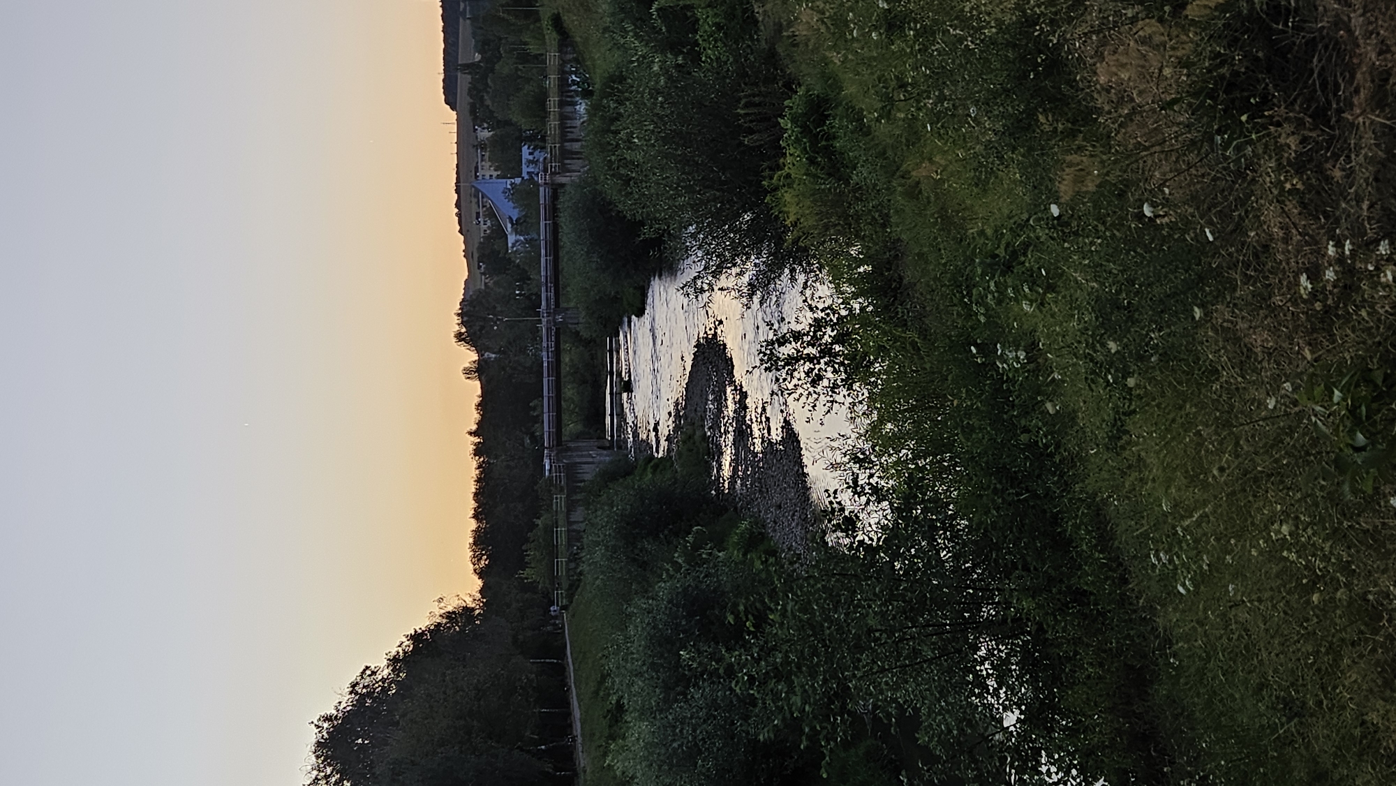The image depicts a scenic view of the Torysa River in Prešov, Slovakia, at dusk. The river, with its shallow, rocky bed and gently flowing water, is the central focus. The banks of the river are densely covered with lush green vegetation, including trees and shrubs, with some scattered wildflowers.
A bridge spans the river in the middle ground, supported by concrete pillars. The bridge appears to be part of a road or pathway, with railings along its sides. On the far bank, beyond the bridge, there is a well-maintained grassy area with a pathway, suggesting a park or recreational zone. In the distance, the cityscape of Prešov is visible, with buildings and trees dotting the horizon. A distinctive blue-tiled building with a sharp, pointed roof stands out among the other structures.
The sky above is a gradient of soft colors, transitioning from a pale yellow near the horizon to a light blue at the top, characteristic of a sunset or sunrise. A faint, small light, possibly a star or planet, is visible in the upper portion of the sky. Two figures are faintly visible walking on the path near the bridge, indicating human presence in the scene. The overall impression is one of tranquility and natural beauty, with a blend of the urban and natural environments of Prešov.