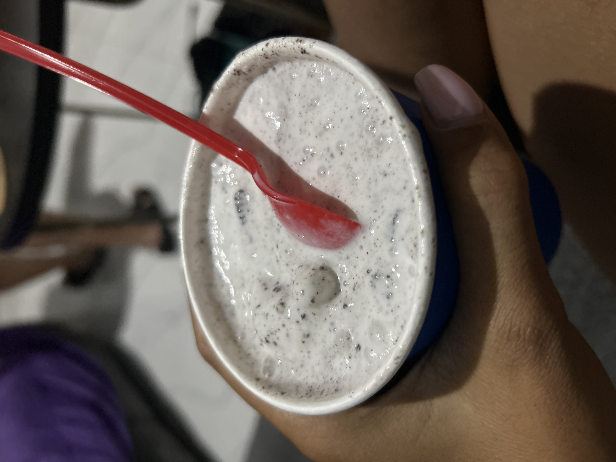 A close-up shot shows a person's hand holding a blue paper cup filled with a white, frothy beverage, likely a milkshake, with dark speckles throughout. A bright red plastic spoon is submerged in the drink. The background is blurred, but suggests an indoor or outdoor seating area with a dark table and hints of people. The image is a mid-shot, focusing on the beverage and the hand.