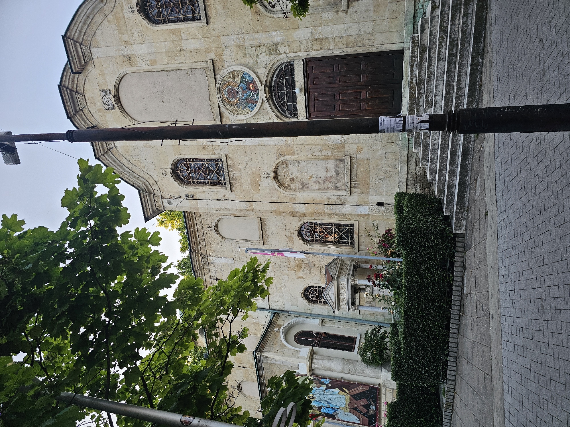 A full shot captures the exterior of a stone church in an urban setting, as viewed from a street level. The church features a weathered stone facade, arched windows with intricate metal grilles, and a large wooden double door. A mosaic of a religious figure is mounted above the entrance. The building is flanked by a neatly trimmed hedge and a flower bed. Paved sidewalks and a cobblestone street lead up to the church steps. A lamppost stands prominently in the foreground, its dark metal pole contrasting with the lighter stone of the church. Lush green tree foliage partially obscures the upper left section of the image. The sky appears overcast.