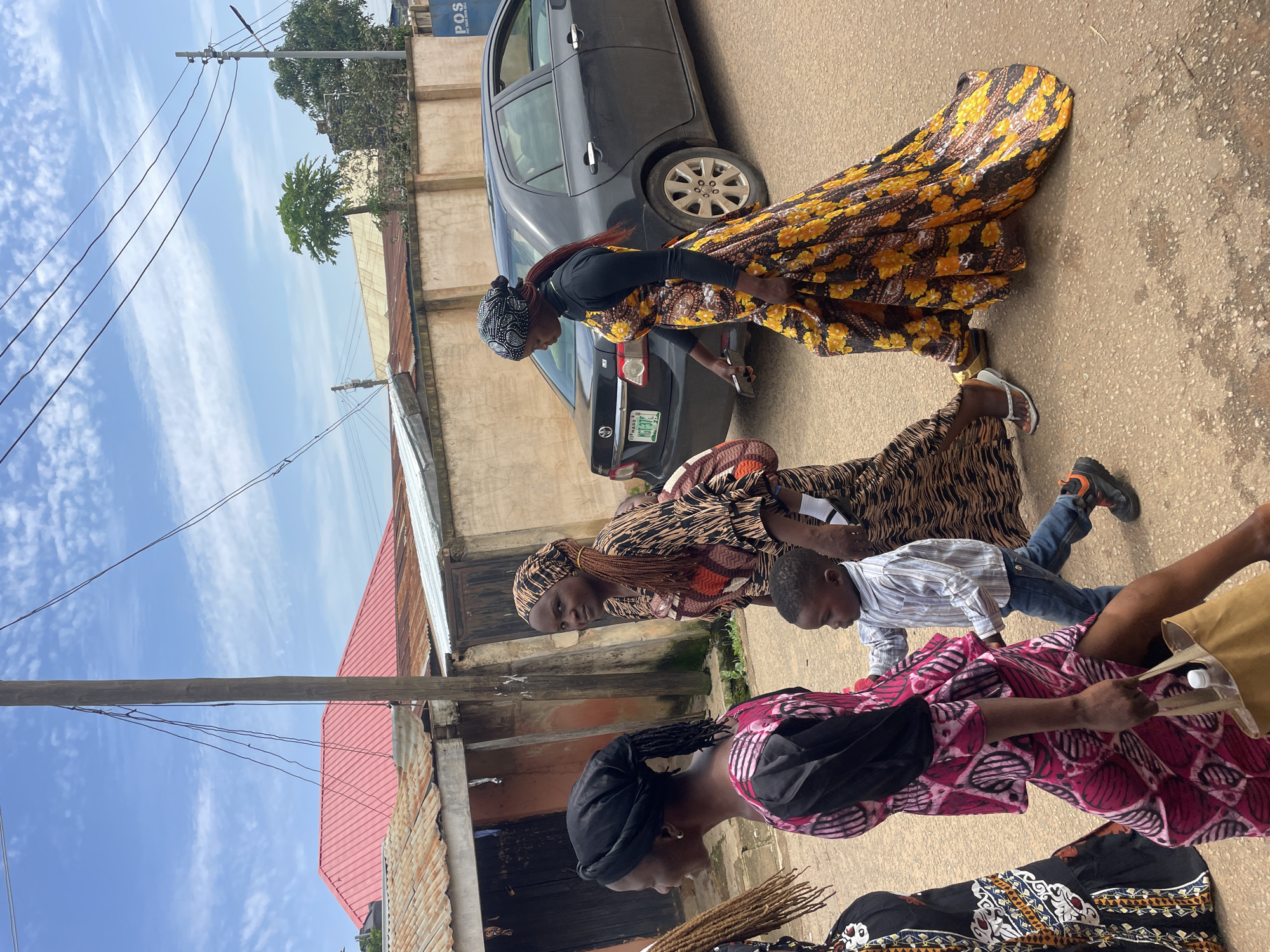 In Jos, Nigeria, on a cloudy day, a group of people, including three women and a young boy, walk along a paved road. The woman on the left wears a black headscarf and a vibrant pink and black patterned dress, carrying a tan bag. The middle woman, with a patterned brown and black headscarf and dress, walks alongside the boy. The boy, dressed in a striped shirt and blue jeans, holds onto her. The woman on the right wears a black long-sleeved shirt under a yellow and orange floral print dress and a black and white patterned headscarf. Behind them, a dark-colored sedan is parked. The background features a mix of buildings with corrugated metal roofs, some appearing weathered, and a few trees. A utility pole with multiple wires stretches across the frame, and a sign for "POS" is visible on a building.