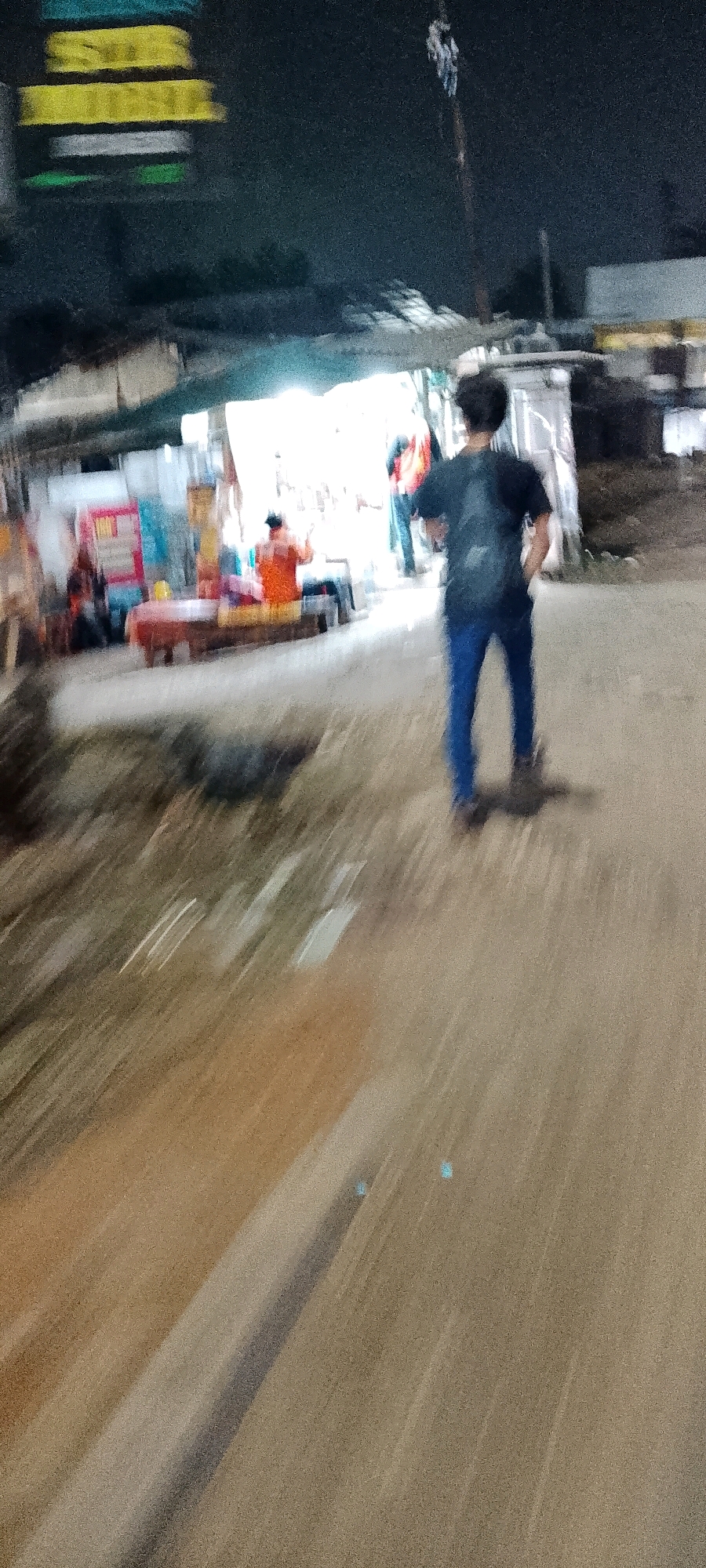 The image shows a dimly lit, outdoor scene at night, likely in Cileungsi, Indonesia. In the foreground, the lower half of the image is heavily blurred, indicating motion. A person, possibly male, is walking away from the camera. They are wearing a dark t-shirt and blue pants, and appear to have a backpack. The ground is a dirt or gravel road with some light-colored streaks, likely from light sources or reflections.
In the midground, there is a slightly more focused area with a market stall or food stand. A person in an orange shirt is visible behind the counter, and there are various colorful items on display. Other figures are present, but they are indistinct due to motion blur and low light. In the background, there are structures that appear to be shops or buildings, with some glowing signs that are also blurred. The overall atmosphere is one of a busy, nocturnal commercial area. The scene suggests a vibrant street life, even in the darkness.