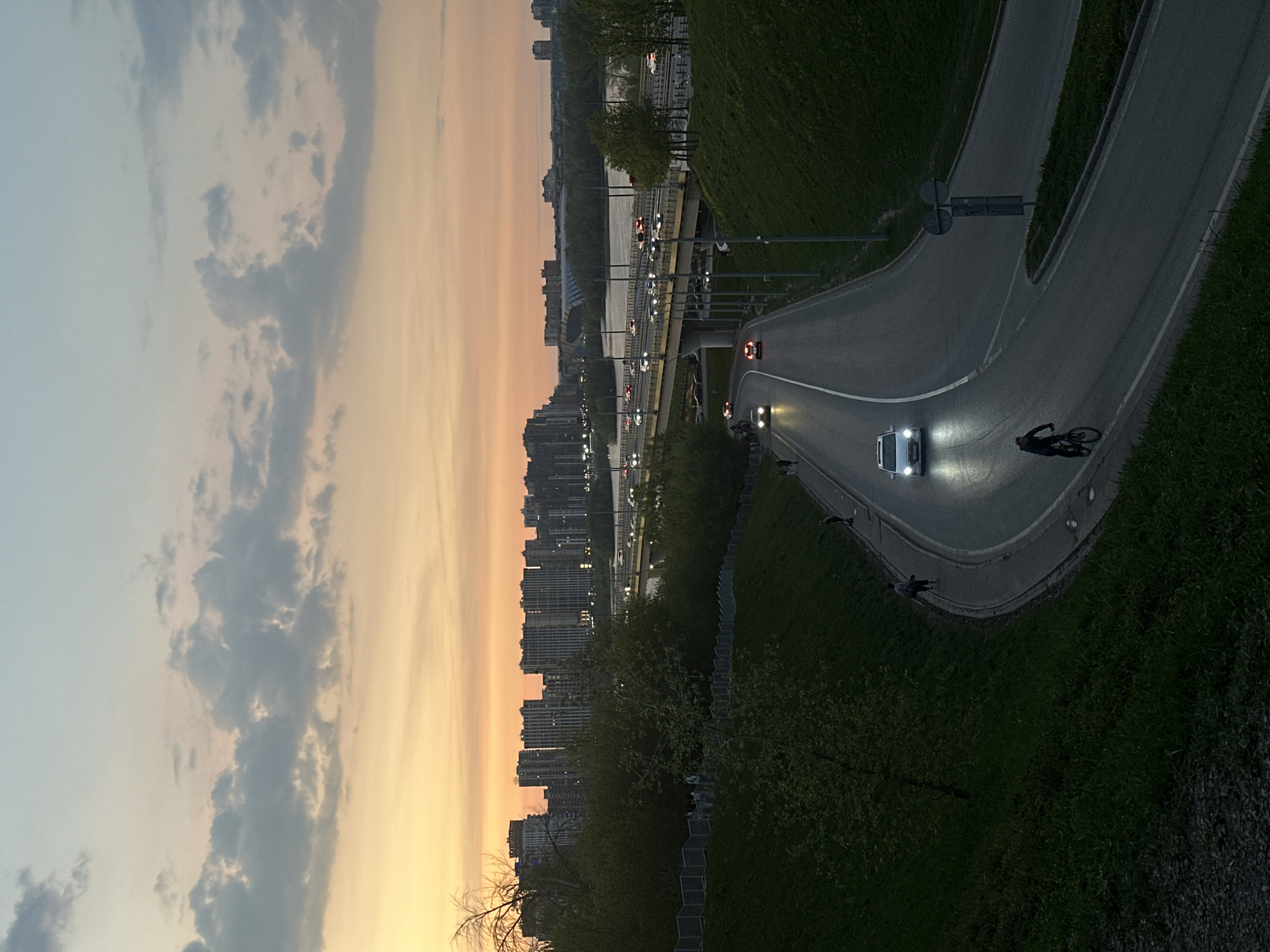 The image presents a view of Kazan, Russia, at dusk. The sky transitions from pale blue to warm orange. The skyline shows numerous tall buildings. A river is spanned by a bridge with moving traffic, indicated by the presence of car headlights. In the foreground, a curved road is visible, with a white car illuminated with headlights and a cyclist. Green vegetation is present on either side of the road.