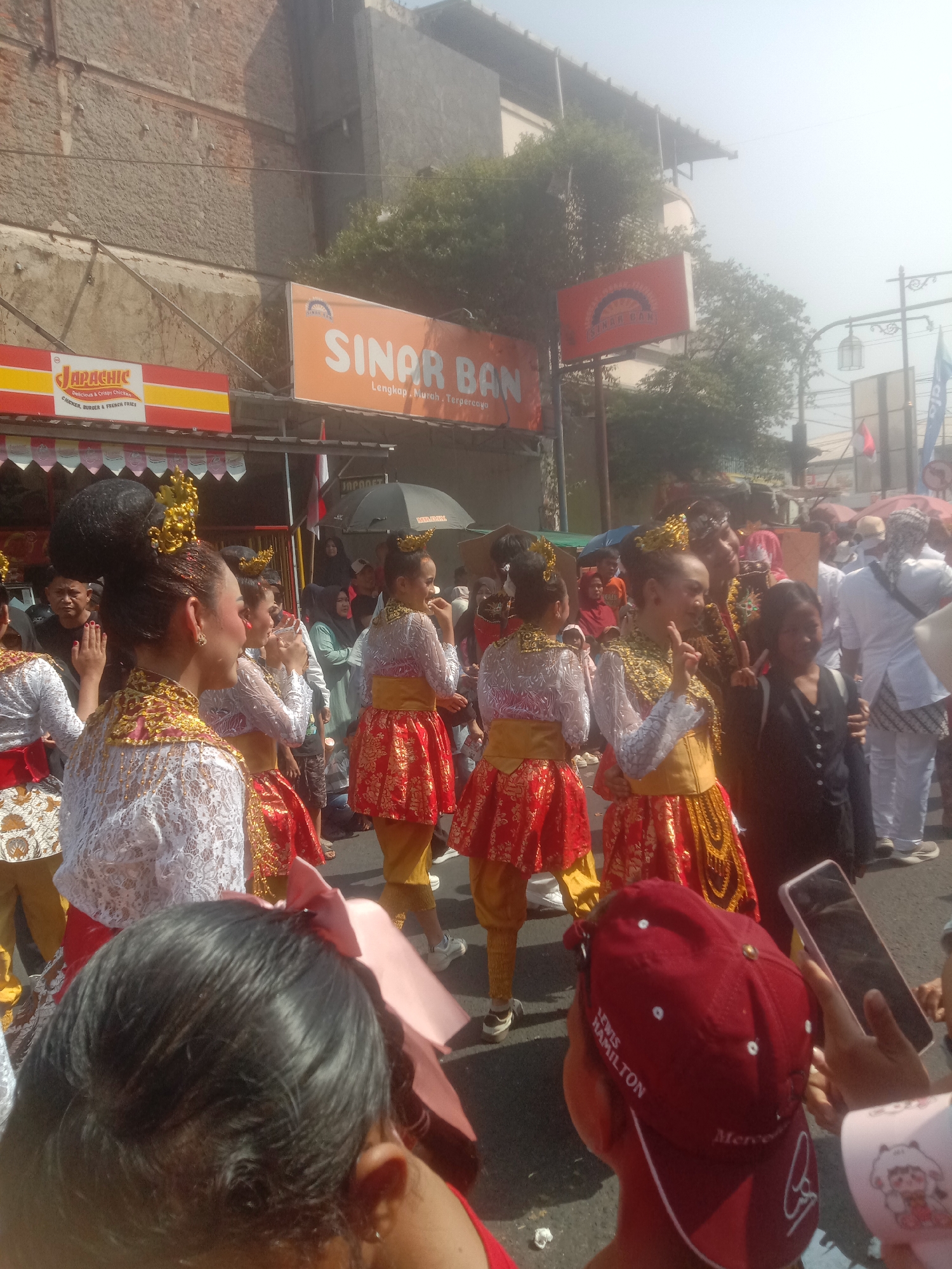 The image captures a lively street scene in Cianjur, Indonesia, likely during a cultural event or parade. In the foreground, several individuals are dressed in traditional vibrant costumes, featuring intricate gold embellishments and red skirts. One person in the center is performing a hand gesture, possibly part of a dance or performance. Behind them, a crowd of onlookers is visible, some holding up their phones to record the event. Signs for "JAPACHIC" and "SINAR BAN" are prominently displayed on buildings along the street, indicating commercial establishments. The environment appears to be outdoors, with a bright, hazy sky suggesting a sunny day. The presence of traditional attire, a gathering of people, and the general atmosphere point towards a cultural celebration in the city of Cianjur.