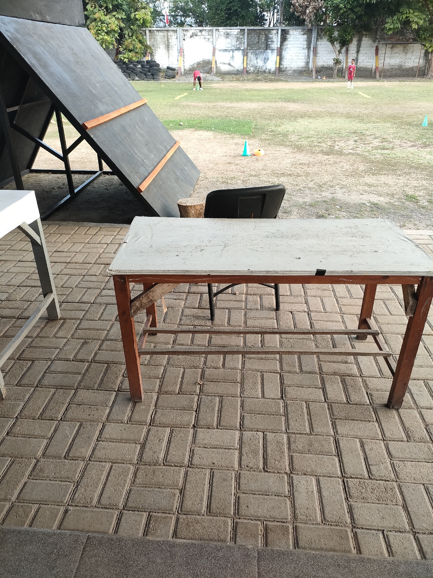 A medium shot captures an outdoor scene in Banguntapan, Indonesia, with a focus on a weathered wooden table in the foreground. The table, exhibiting a light-colored, textured surface and brown wooden legs, sits on a brick-paved area. To the left of the table, a portion of a white table is visible. Behind the wooden table, a black chair is partially obscured. In the background, a large, dark inclined obstacle structure dominates the left side of the frame. Beyond this, a grassy field stretches out, marked with some orange and blue cones. Several individuals are present on the field, including one person in a red shirt bending down, and another in a red shirt standing further back. A white wall with some yellow and red accents runs along the edge of the field, with trees and foliage visible behind it. The overall impression is of an outdoor training or recreational area.