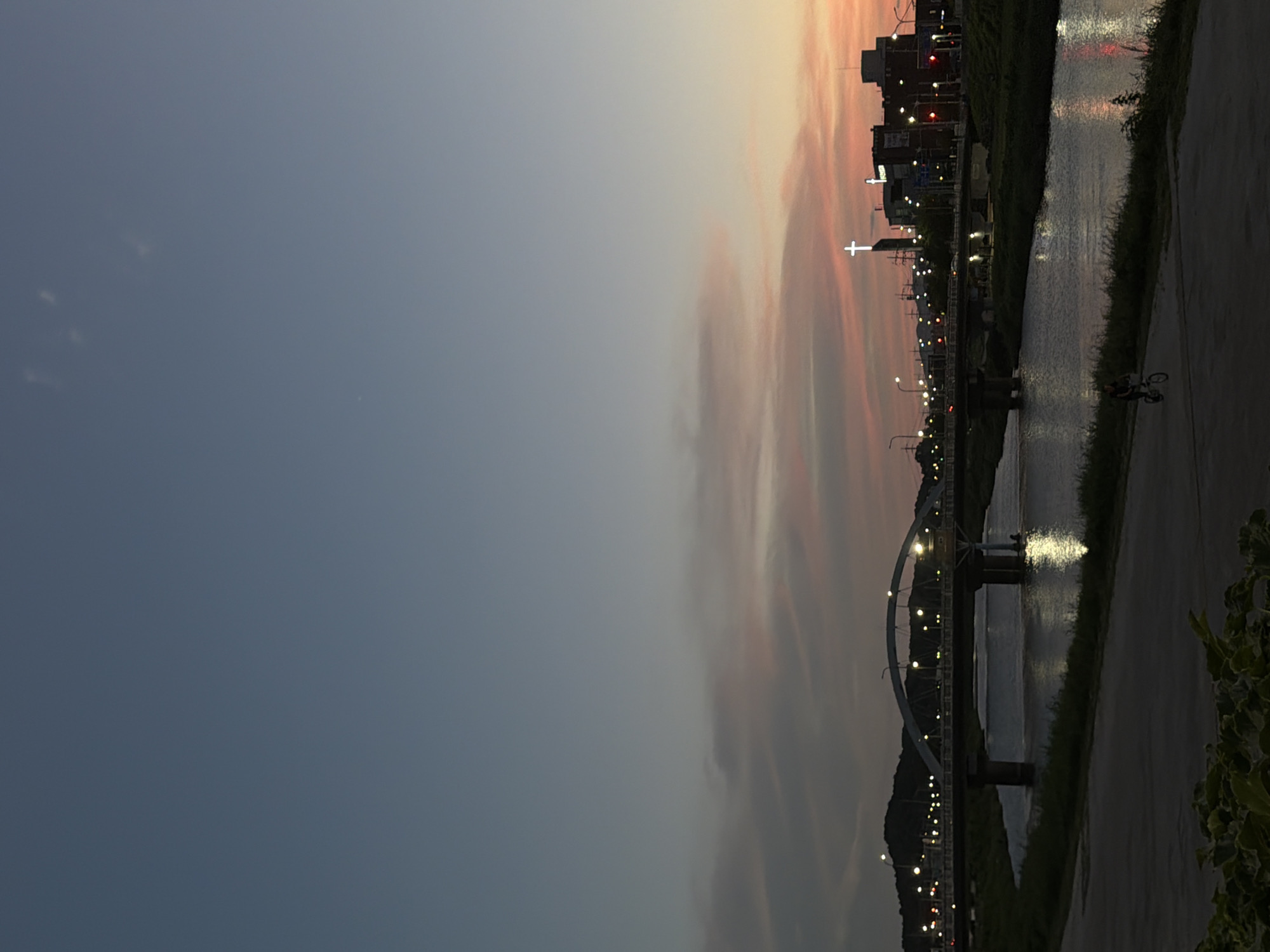 An urban landscape at dusk in Dongducheon-si, South Korea, features a river reflecting the soft glow of streetlights and a twilight sky painted with hues of pink and orange. A bridge spans the river, its arches illuminated by numerous lights. On the far bank, buildings are visible, including a prominent church steeple topped with a glowing cross. The foreground shows a paved path alongside the river, where a lone cyclist is riding. Lush green foliage lines the riverbank in the lower left of the frame.