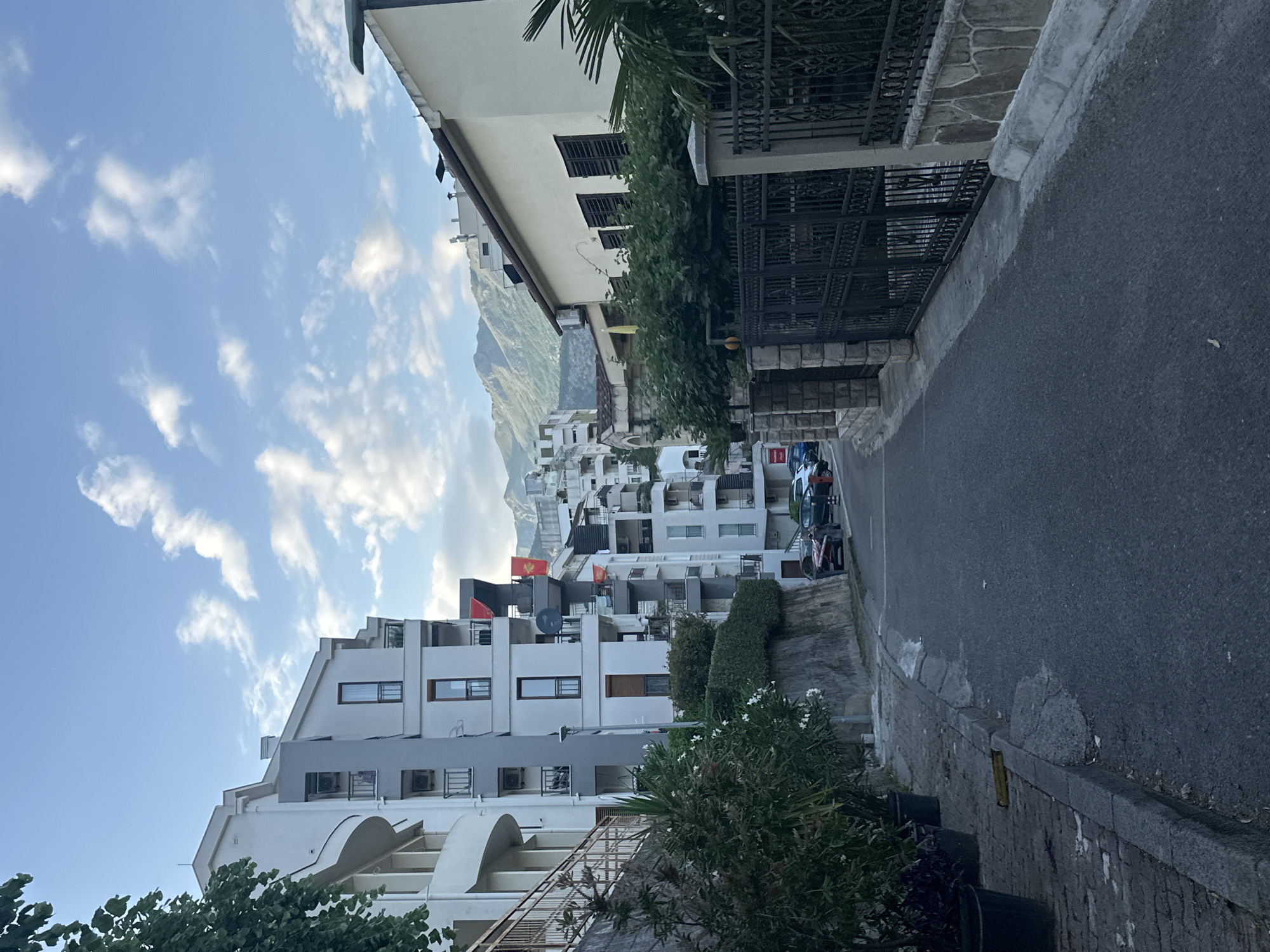 The photograph depicts a street scene in Budva, Montenegro. The street is narrow and paved with asphalt. Buildings line both sides of the street, primarily multi-story residential buildings with white facades and balconies. Some buildings have Montenegrin flags displayed. A mountain range is visible in the background. The sky is light blue with scattered clouds. Several parked vehicles are seen along the side of the street. The scene is generally well-lit, suggesting it is daytime. Vegetation, including trees and bushes, are present along the street.