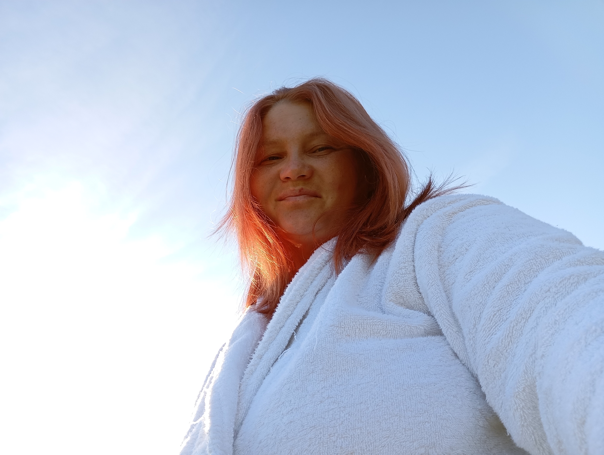 A woman with reddish-pink hair and fair skin is captured from a low angle, looking upwards. She is wearing a fluffy white bathrobe. The background is a clear blue sky with bright sunlight creating a lens flare effect on the left side of the image. The lighting suggests it is daytime, possibly early morning or late afternoon, due to the warmth of the sun. The image appears to be a selfie taken outdoors. The location is unknown.