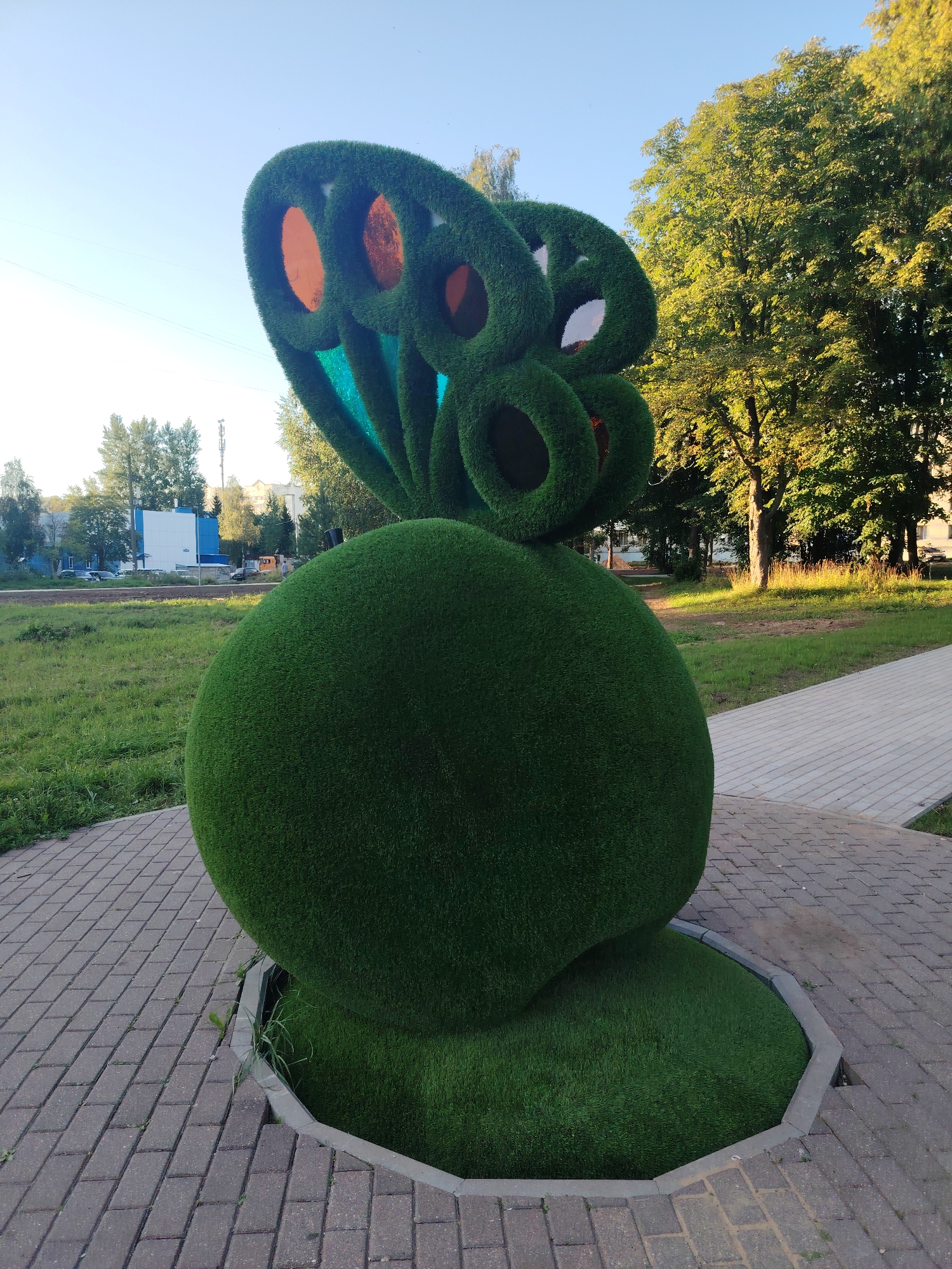 A topiary sculpture resembling a large, stylized butterfly is situated on a brick-paved pathway in a park-like setting. The sculpture consists of a large, spherical green body covered in artificial grass and a set of green, ring-shaped wings also covered in artificial grass. The wings have large circular openings, some of which reveal an orange-colored material beneath. The sculpture is surrounded by a low, hexagonal concrete border, within which artificial grass also grows. The pathway is constructed of grey and reddish-brown bricks arranged in a patterned layout. Beyond the pathway and sculpture, there is a grassy area with scattered trees, some of which are illuminated by sunlight. In the background, buildings and a cell tower are visible, suggesting an urban or suburban environment. The sky is clear and blue, indicating fair weather. The scene appears to be part of a public park or outdoor recreational area in the city of Konakovo, Russia. There are no people or animals visible in the image.