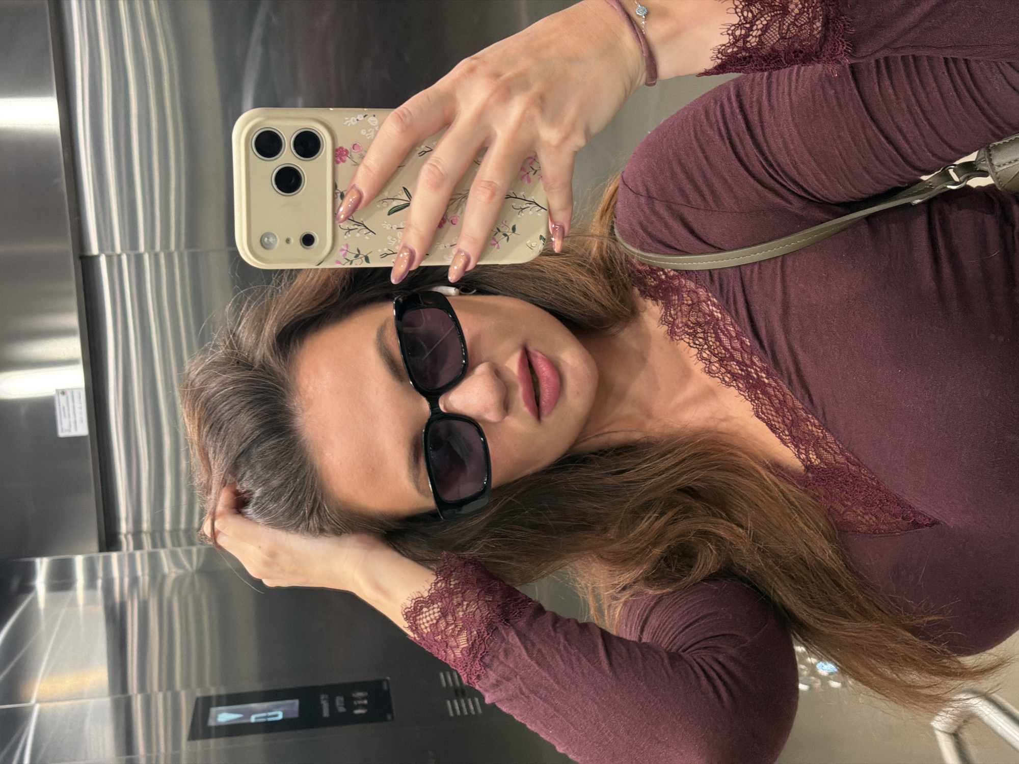 A brunette woman is taking a selfie in what appears to be an elevator in Belgrade, Serbia. She is wearing sunglasses and a dark purple long-sleeved shirt with lace detailing. She is holding a smartphone in her right hand, which has a floral phone case and manicured nails. Her left hand is partially visible, touching her hair. The elevator has a stainless steel interior and a control panel is visible.