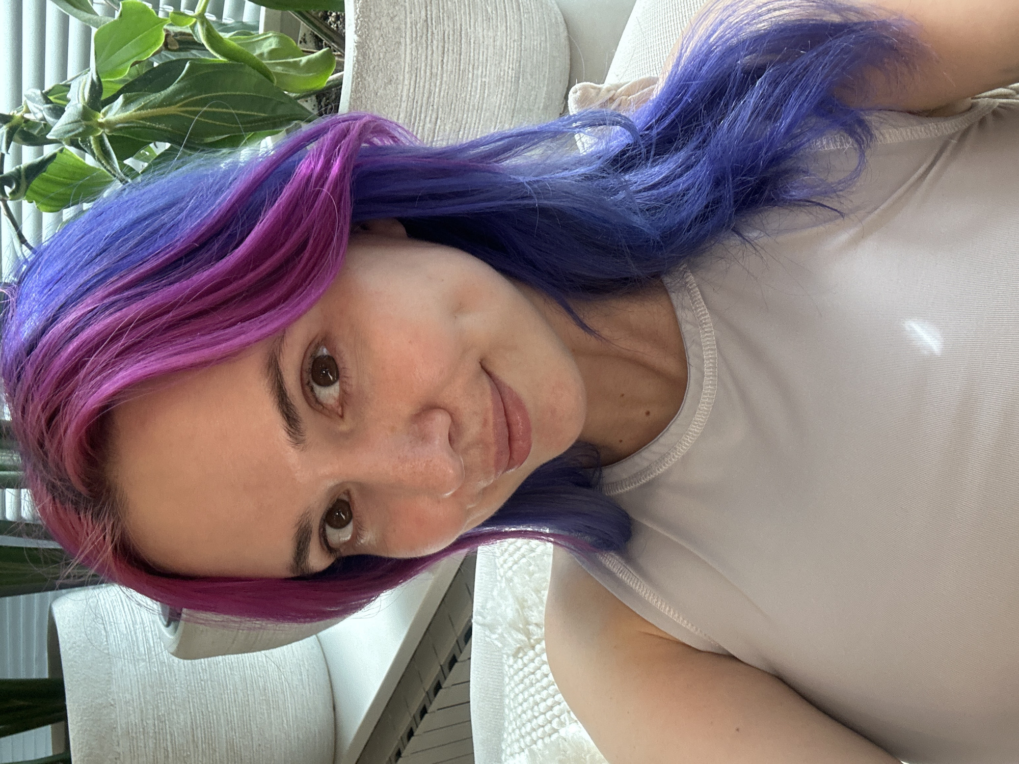 This media file presents a close-up, tilted portrait of a woman indoors in the City of Zagreb, Croatia. The woman, likely in her 20s or 30s, looks directly at the camera with a gentle smile. Her most distinctive feature is her vibrant, multi-colored hair, which transitions from bright pink and magenta tones near her face and scalp to a deep, vivid blue further back. Her hair is long and appears to be styled loosely. She has fair skin with some visible freckles and warm brown eyes. She is wearing a plain, light-colored, possibly off-white or light grey, sleeveless top.
The setting appears to be a brightly lit room during the daytime, with natural light falling on her face and clothing. In the background, a green, leafy houseplant is visible, suggesting a natural element within the indoor environment. A light-colored, textured piece of furniture, possibly a sofa or armchair, also occupies a portion of the background, indicating a comfortable and relaxed domestic setting. There is no visible text in the image.