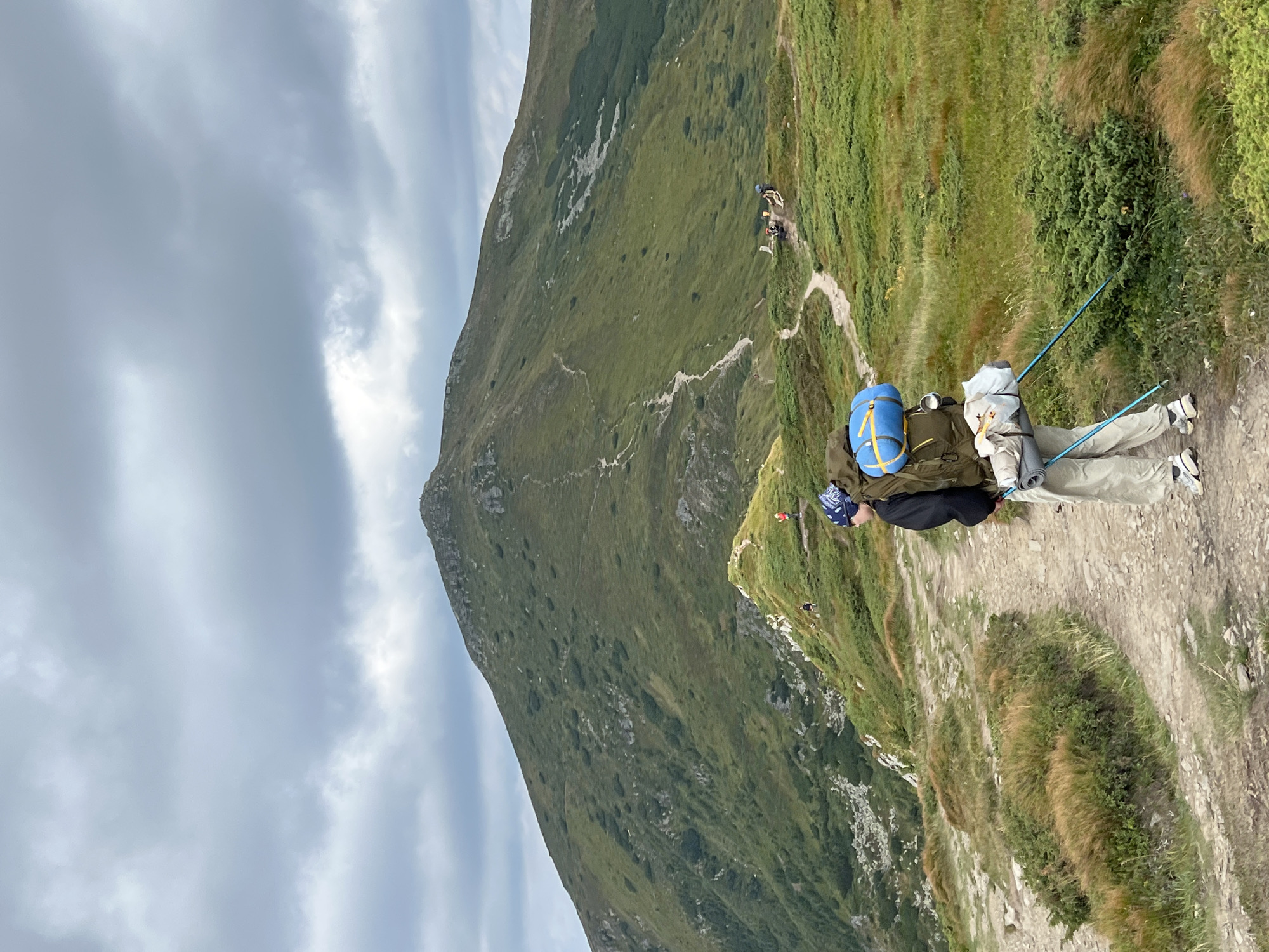 The image shows a mountain landscape in Luhy, Ukraine. In the foreground, a person is hiking on a trail, carrying a large backpack and using trekking poles. The person is wearing a bandana, a dark jacket, and light-colored pants. The trail winds through grassy hills covered in low-lying vegetation and scattered rocks. In the background, a large, green mountain dominates the scene, with a winding path visible on its slope. The sky is cloudy with a mix of gray and white clouds. Further in the distance, on a ridge to the right, a few other hikers are visible, some sitting and resting. The overall scene depicts a remote, natural environment suitable for outdoor recreation.