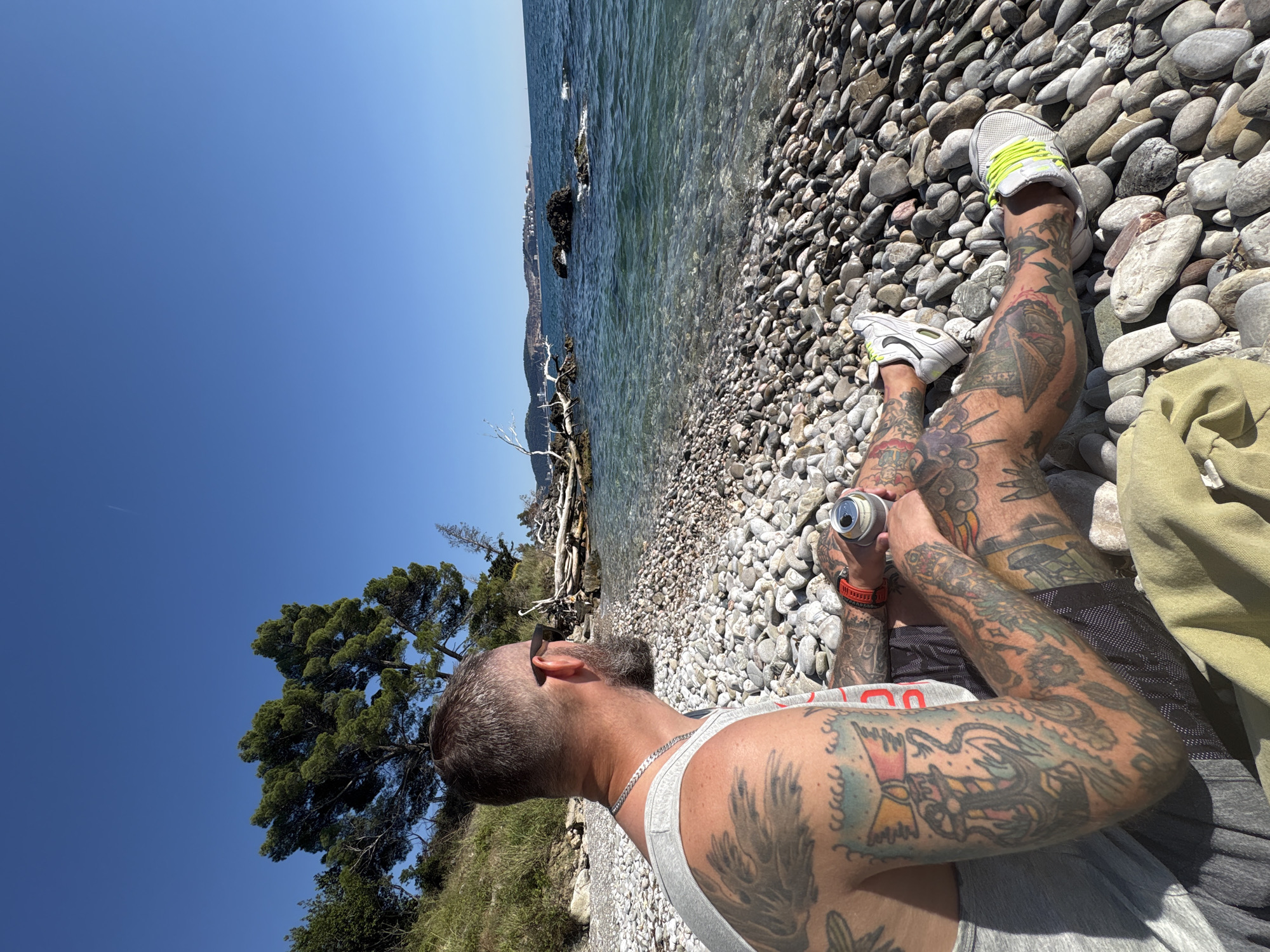 A man with tattoos sits on a pebble beach in Bar, Montenegro. The man has short, dark hair and a beard, and he is wearing sunglasses, a grey tank top, and shorts. He is holding a beverage can in his hands. His legs are crossed. He is wearing white sneakers with neon yellow laces. The beach is covered in grey pebbles of varying sizes. Behind him is clear water, and a coastline is visible in the distance. A small tree is to his left. The sky is a clear blue.
