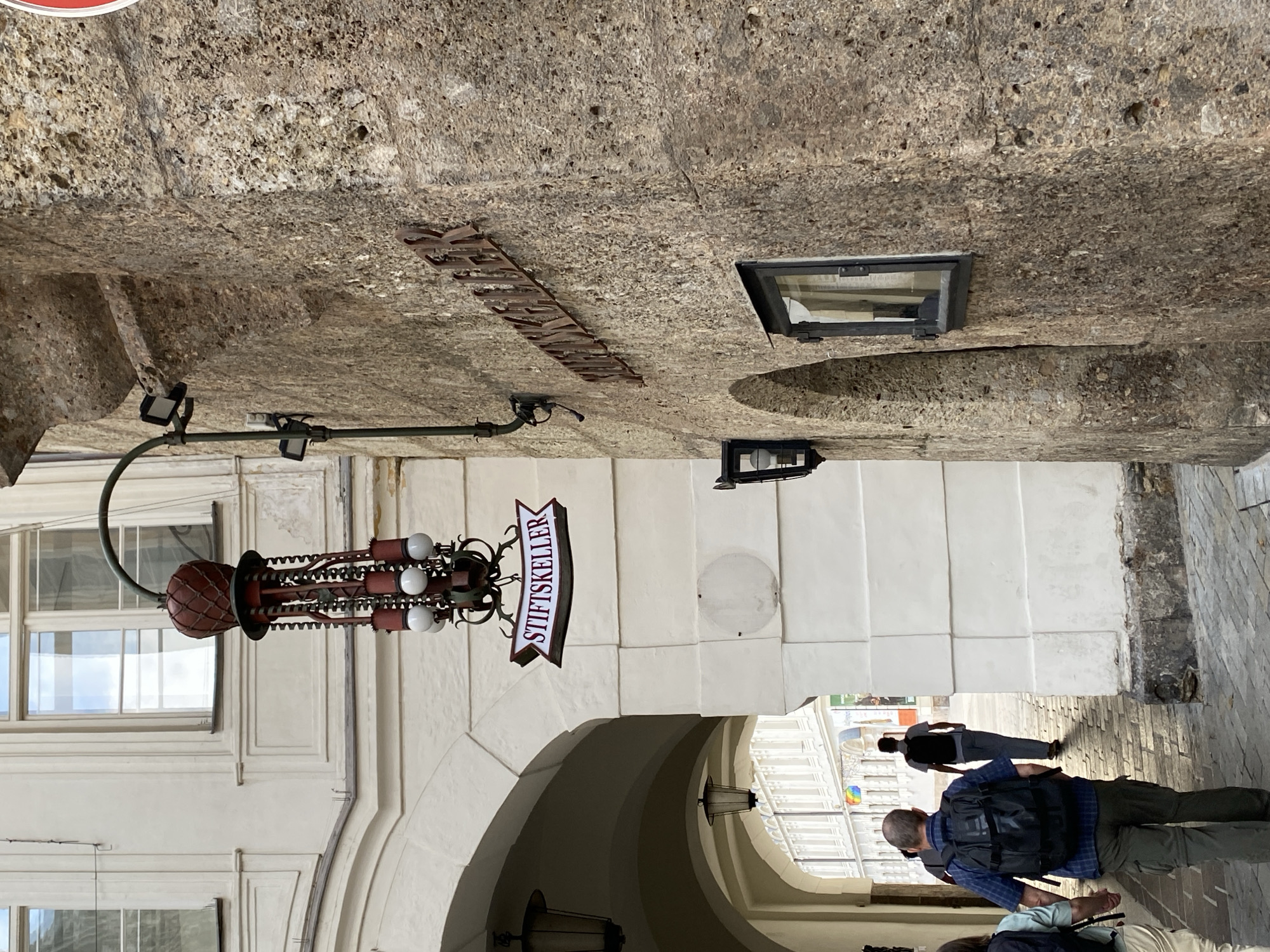 The image shows a street scene in Innsbruck, Austria, featuring historical architecture and pedestrian activity. A man with a backpack is walking under an archway, followed by another person. To the right, a stone building with a rough-hewn facade displays a wrought-iron sign that reads "STIFTSKELLER". Above the sign, a decorative light fixture with red and white elements hangs from an arm attached to the building. Across the archway, a white building with large windows is visible. The ground is paved with cobblestones. The overall impression is of a charming, old-world European street.