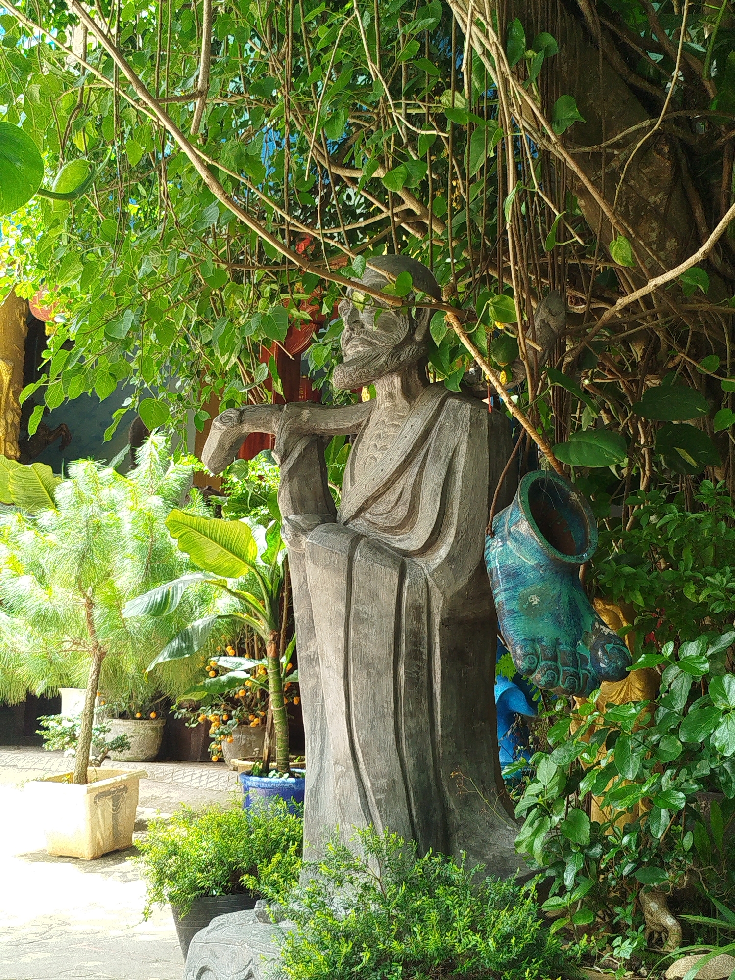 A wooden statue of a robed figure with a beard stands outdoors, partially obscured by dense green foliage and hanging vines. The statue holds an object across its chest with its left arm. To the right of the statue, a turquoise, foot-shaped ceramic object hangs from a string. The statue and decorations are situated in a lush, garden-like setting, with various potted plants visible in the background, including a small pine tree and banana plants with yellow fruit. In the far left, a glimpse of a yellow structure and a blue wall can be seen. The scene suggests an outdoor space in Da Lat, Vietnam, likely a garden or a temple grounds, given the presence of statues and spiritual iconography.