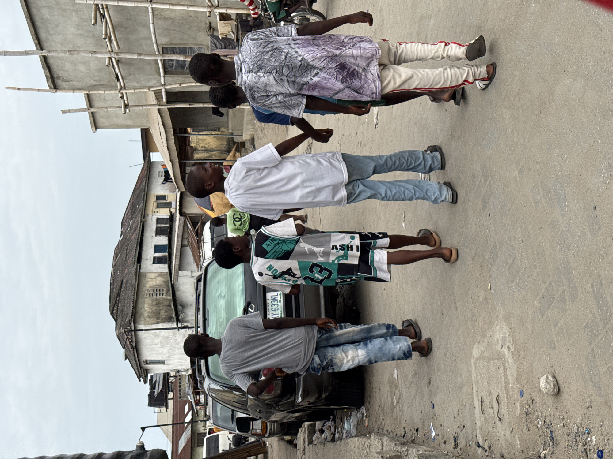 This image captures a daytime street scene in an urban area, likely Lekki, Lagos, Nigeria, under an overcast sky. Several young males are standing on a paved road, which appears somewhat worn with scattered debris like small rocks and plastic caps.
In the foreground, a young male in a light grey t-shirt and faded blue jeans stands by the rear of a black car, holding an orange object, possibly fruit. To his right, another male wears a distinctive multi-patterned white, black, and teal shirt with shorts and sandals; text like "AIR", "ASH", and "NO" is visible on his attire. The black car, possibly an SUV or minivan, displays a Nigerian license plate that reads "LAGOS KY-633ML".
Further down the street, a young male in a white t-shirt and blue jeans looks upwards. Behind him, two other individuals are partly visible: one in a purple and white patterned shirt with light-colored trousers, and another in a blue shirt. A person wearing a green shirt with a visible "CHANEL" logo is partially obscured behind others.
The background features several buildings of varying styles and conditions. One prominent concrete building is surrounded by bamboo scaffolding, suggesting ongoing construction or renovation. The overall environment indicates a busy, lived-in community. The expressions of the individuals are generally relaxed, and they appear to be casually gathered on the street.