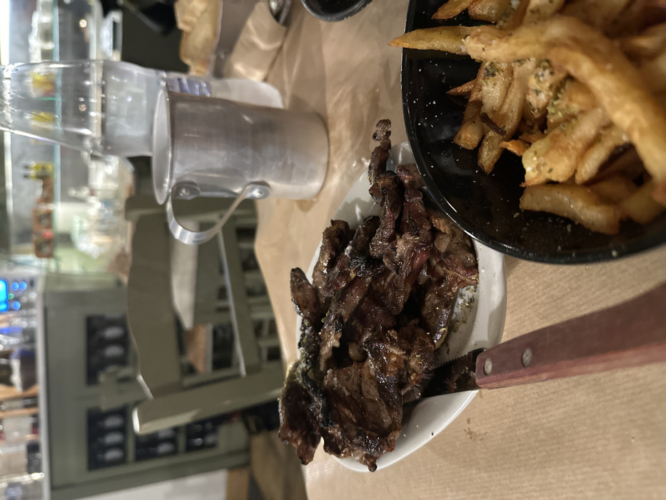 A close-up, slightly rotated view captures a casual dining experience, likely at a restaurant in Nea Erithrea, Greece. The primary subjects are two dishes: a generous serving of grilled meat and a bowl of french fries.
The grilled meat, appearing to be thinly sliced or small cuts, is dark brown with visible charring and glistening fat, suggesting it's freshly cooked and juicy. It's seasoned with coarse salt and possibly herbs, presented on an oval white plate. A steak knife with a wooden handle and a serrated blade is partially submerged into the meat, indicating it's ready to be eaten.
Adjacent to the meat is a black bowl filled with golden-brown french fries. They are thick-cut, lightly seasoned with salt and what appears to be dried herbs, possibly oregano, a common seasoning in Greek cuisine.
In the upper left of the frame (when the image is correctly oriented), a silver-colored metal pitcher with a handle is visible, likely holding water or another beverage. The table beneath the dishes is covered with light brown paper, often used in casual eateries to line tables.
The background is softly blurred, showing an indoor restaurant environment. Distant, out-of-focus elements suggest a bar area or shelving with various bottles and glasses. A distinct blurry blue light source, possibly from an electronic display or sign, adds to the evening ambiance. There are no people visible, and no discernible text appears in the image. The artificial lighting suggests the meal is taking place in the evening or at night.