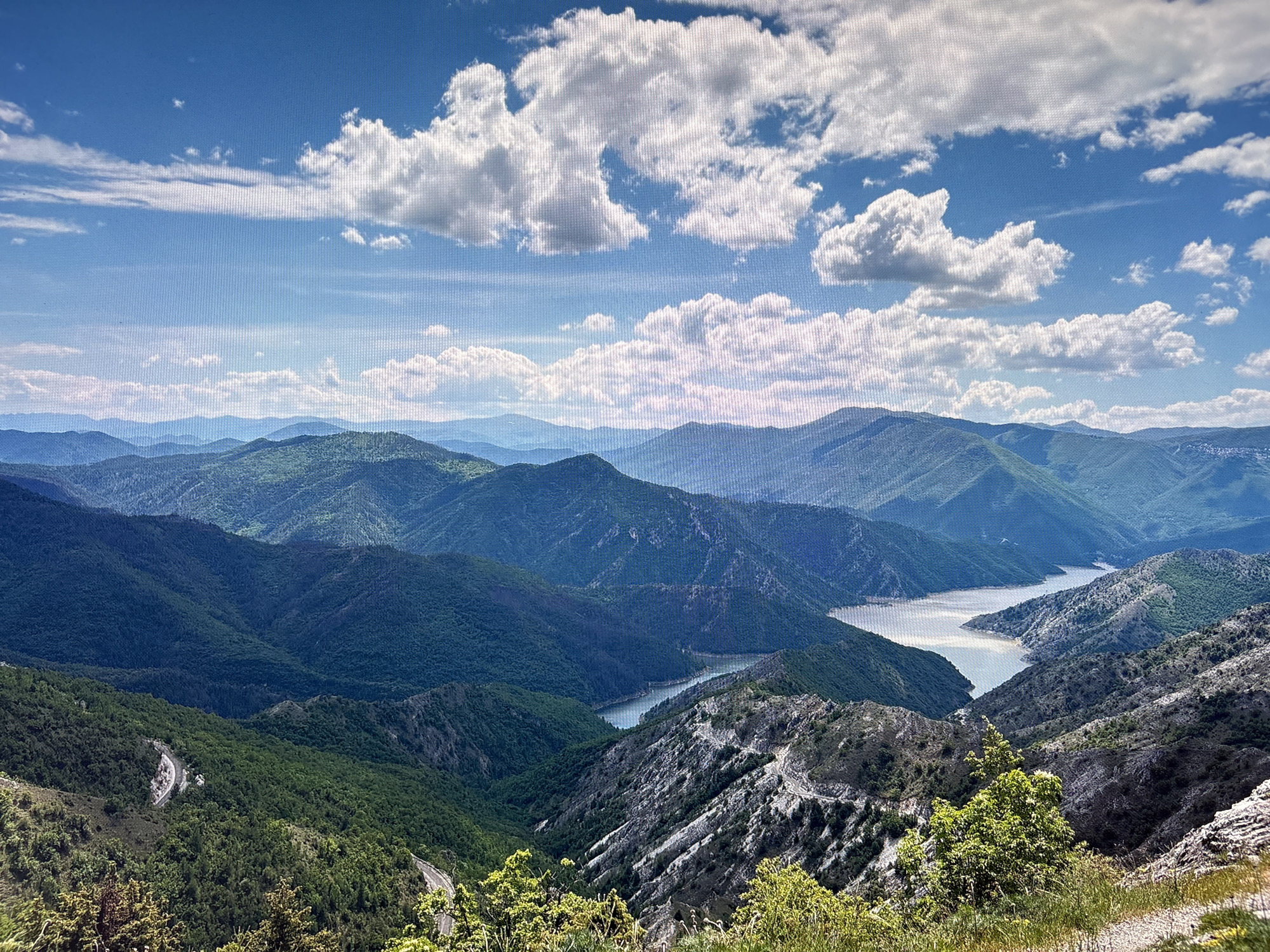 The image shows a landscape view with a chain of green mountains fading into the distance under a blue sky with scattered white clouds. A body of water, likely a river or lake, winds through the mountains. A winding road is visible on the lower left side of the image. The location and city are unknown.