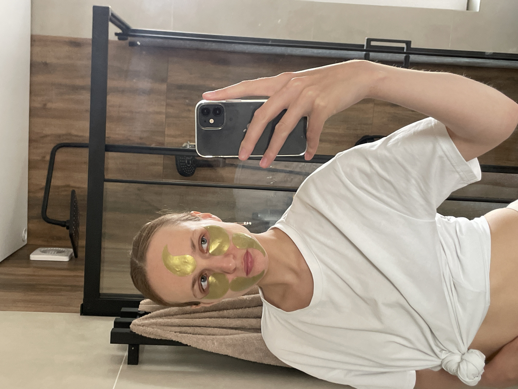 A young woman is taking a selfie in a bathroom. She is wearing a white t-shirt tied in a knot at the waist and has gold-colored eye masks on her face. She is holding a black smartphone in her right hand. The bathroom has tile flooring and wooden paneling on the walls. A black metal shower frame is visible in the background, along with a towel rack with a beige towel on it. The location is Podshivalovo, Russia.