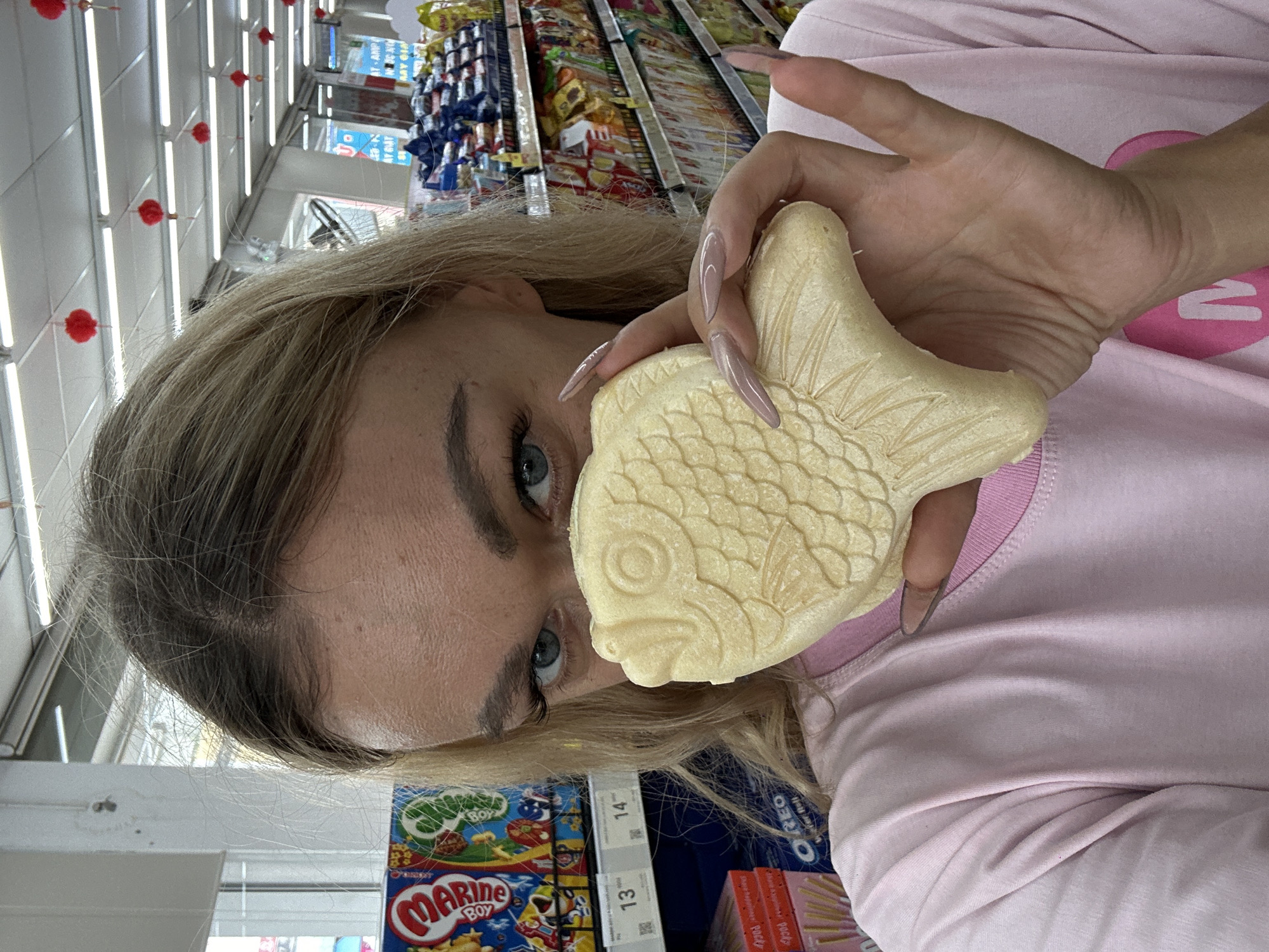 A fair-skinned person with blonde hair and blue eyes is holding a fish-shaped pastry (Taiyaki) in front of their face, appearing to be inside a supermarket or convenience store. The person is wearing a light pink long-sleeve shirt. The scene suggests an Asian food store, as various snack items are visible on shelves in the background. There is no information available to determine if this photo was taken in Đồng Nai, Vietnam.