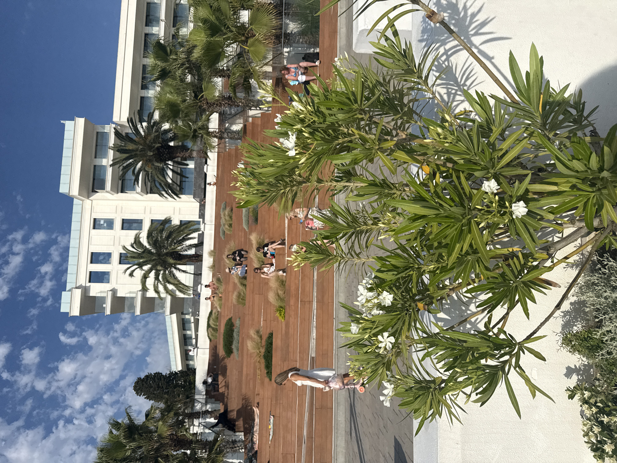 This media file depicts a sunny day in Sochi, Russia, featuring a modern hotel building, palm trees, and an outdoor tiered seating area made of wood. Several people are visible relaxing on the tiered seating, some sunbathing, while others are engaged in conversation. A woman on a scooter is also present in the foreground. The scene suggests a leisure or vacation setting, possibly a hotel resort or a public space with a view. The weather appears to be warm and clear, with a bright blue sky and scattered clouds. The lush green foliage of the palm trees and other plants indicates a temperate or subtropical climate. The architecture of the hotel is contemporary, with multiple floors and balconies. The overall mood is relaxed and serene, with people enjoying the outdoor environment.