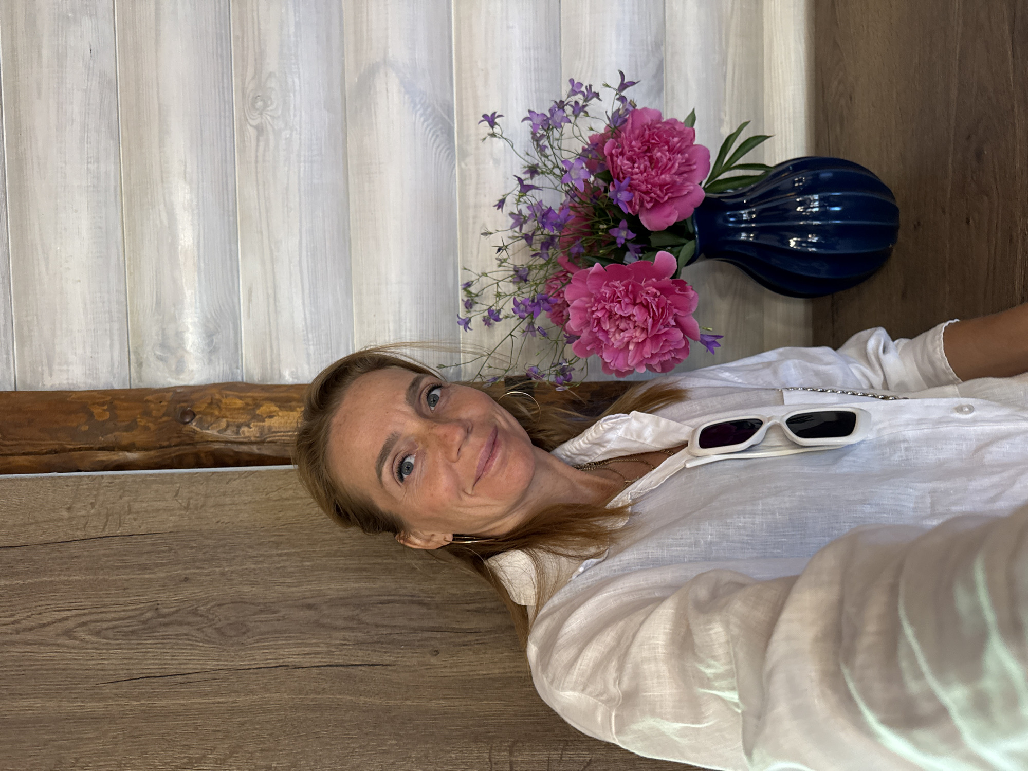 A medium shot shows a middle-aged Caucasian woman with fair skin and freckles posing for a selfie. She has blonde hair pulled back loosely and is wearing a white linen shirt. She has blue eyes and is smiling. A bouquet of pink peonies and small purple flowers in a dark blue vase is to her right. The background features a wooden wall and a horizontally striped light-colored wall. The scene is indoors, likely a cafe or restaurant in Veliky Novgorod, Russia, indicated by the wooden architectural elements commonly found in traditional Russian establishments.
