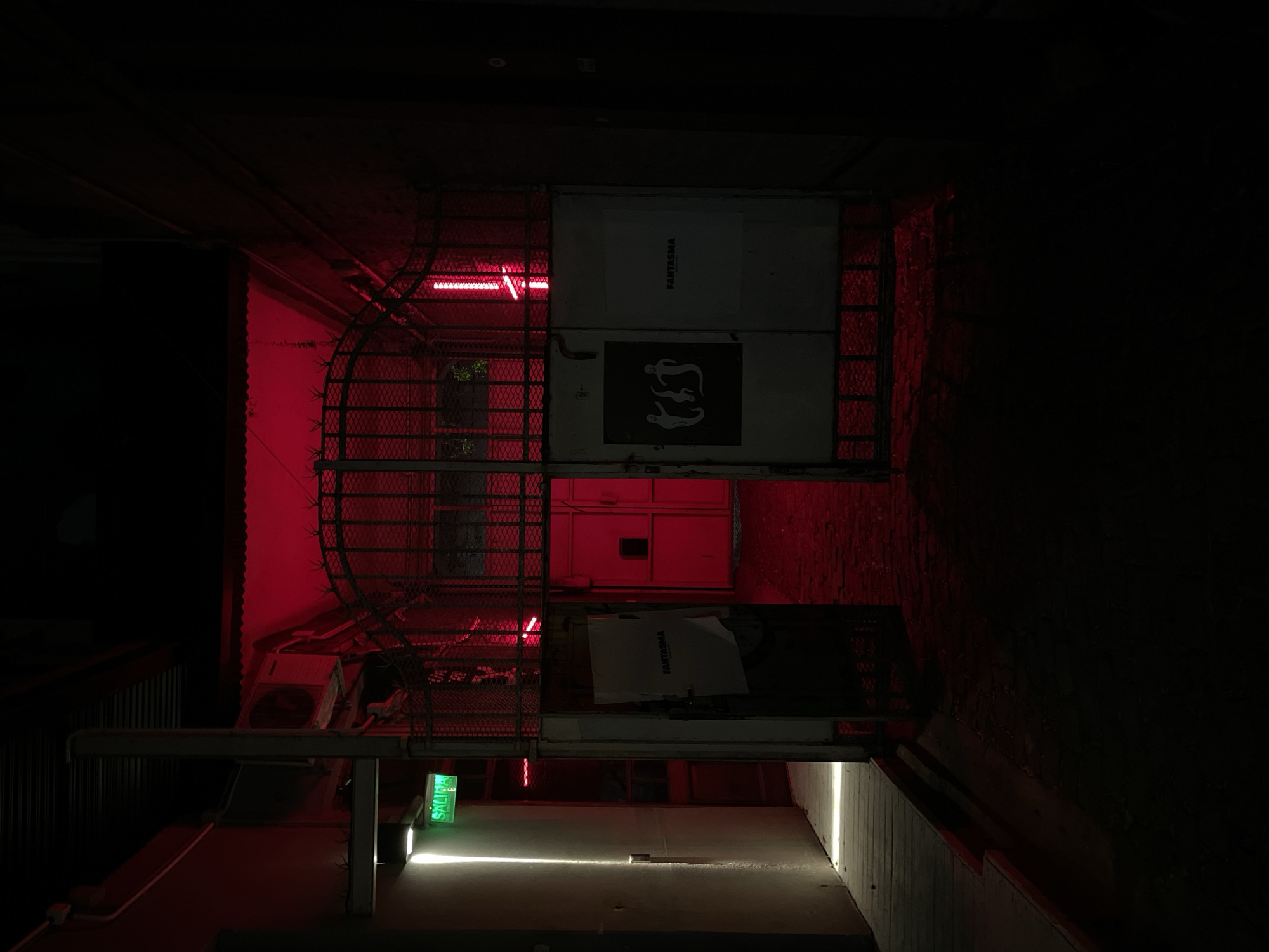 The image displays a dimly lit, narrow passage or interior space, primarily illuminated by striking red light, creating an intense atmosphere. No people are visible.
On the left side of the frame, a tall metal grating or fence structure is prominent, bathed in the red glow from several horizontal neon-like lights running along its length. To the right of this structure, a large, light-colored panel or door displays a black sign. This sign features the word "FANTASMA" (Spanish for "ghost") written vertically, above three stylized white ghost figures.
In the lower left section, partially obscured, is an air conditioning unit. Below it, a bright green rectangular sign, oriented sideways, clearly displays the Spanish word "SALIDA" (EXIT). A strong horizontal line of white light extends from the bottom of this sign across the concrete floor or pathway, suggesting an exit or entrance point. The surrounding walls are dark, appearing industrial or utilitarian, with some textured brickwork visible on the far right.
The scene suggests an urban setting at night, possibly an alleyway, a building's service area, or a club entrance, evoking a mysterious and edgy mood. The visible text, "FANTASMA" and "SALIDA", provides specific location cues, consistent with an establishment in a Spanish-speaking city like Buenos Aires, Argentina. No overt activity is taking place.