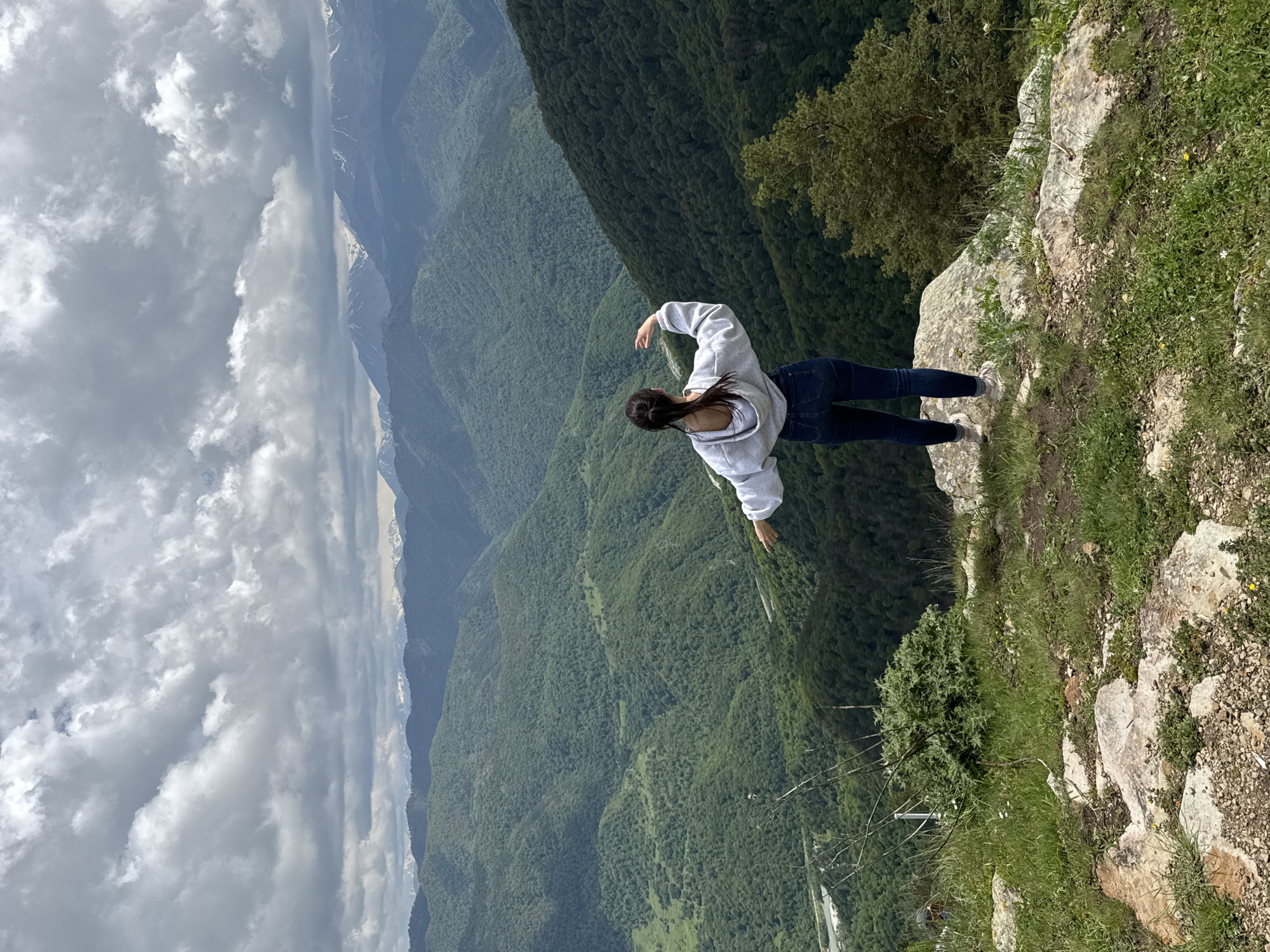 A young woman stands on the edge of a rocky cliff overlooking a vast, green valley filled with rolling mountains. The woman is seen from behind, wearing a grey hoodie and blue jeans, with her arms outstretched as if embracing the expansive landscape. The sky above is partly cloudy, with dramatic grey clouds interspersed with patches of blue, hinting at a dynamic weather system. In the distance, snow-capped mountains form a majestic backdrop. The foreground is characterized by rugged terrain with scattered rocks and green grass. The image captures a moment of solitude and awe in a breathtaking natural setting, potentially in the vicinity of Nizhnyaya Teberda, Russia, a region known for its stunning mountain scenery.