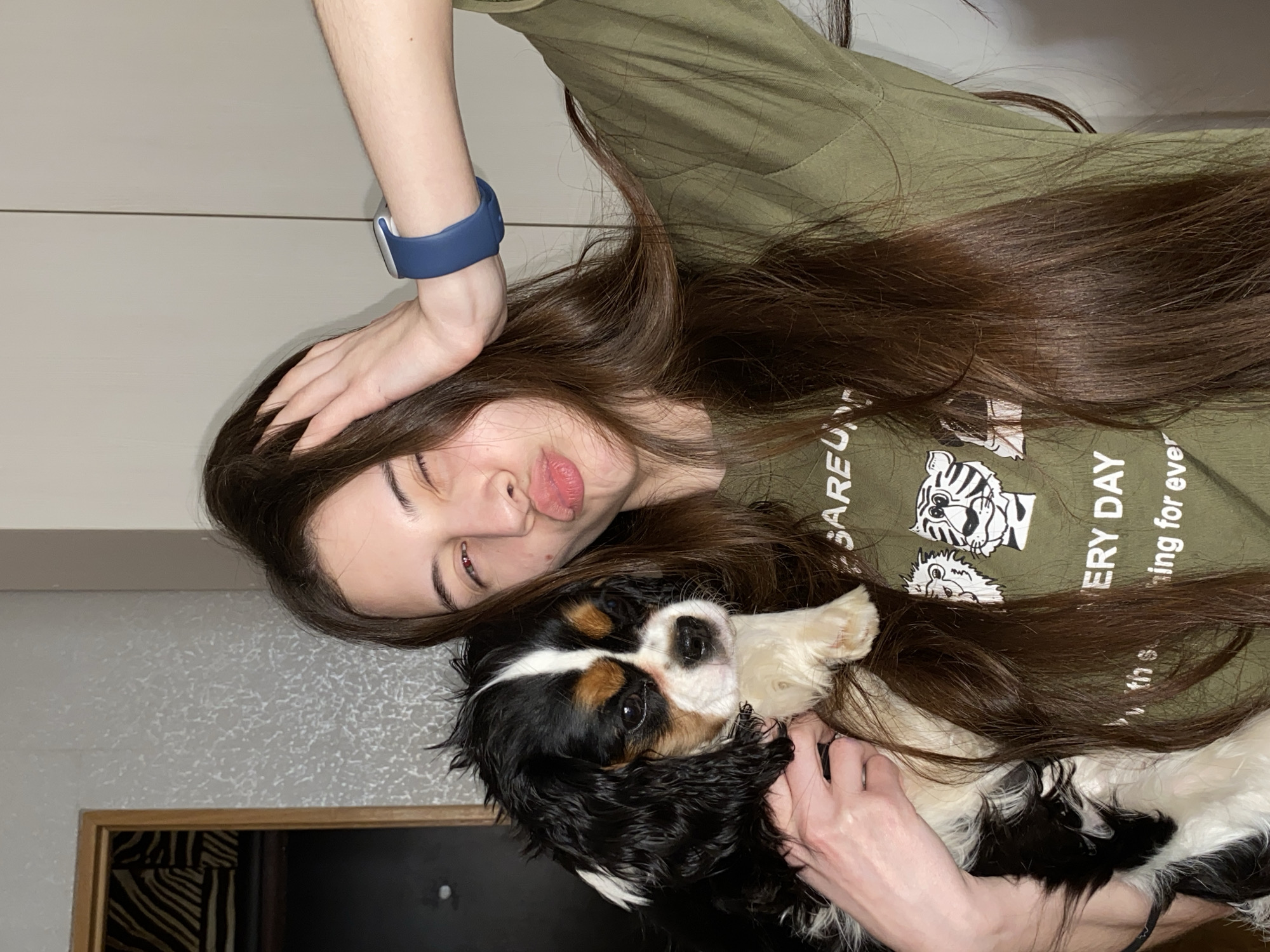The image displays a young woman and a Tricolor Cavalier King Charles Spaniel, captured indoors in what appears to be a home setting. The photo is taken from a slightly elevated angle, with the subjects lying down.
The woman has long, dark brown hair and wears an olive green t-shirt with a graphic print. Her right hand is placed on her head, while a blue smartwatch adorns her left wrist. She is posing playfully, with her lips puckered in a kissy face and her eyes partially closed, conveying a sense of lightheartedness.
Beside her, the dog, a Tricolor Cavalier King Charles Spaniel, features distinct black, white, and tan markings. It is nestled close to the woman, looking towards the camera with dark, soulful eyes. The interaction between the woman and the dog appears affectionate and relaxed.
The background consists primarily of a light-colored wall. In the lower left corner, a glimpse of a darker, possibly textured wall section is visible, alongside a wooden frame bordering a dark opening that reveals a zebra print pattern.
On the woman's t-shirt, parts of the text "EVERY DAY" are clearly visible, with additional words like "AREFUL BALANCING ACT" partially seen. Above this text, two cartoon tiger head graphics are printed. The overall emotion conveyed is one of warmth and playful companionship. This image is contextualized as being taken in Votkinsk, Russia.
