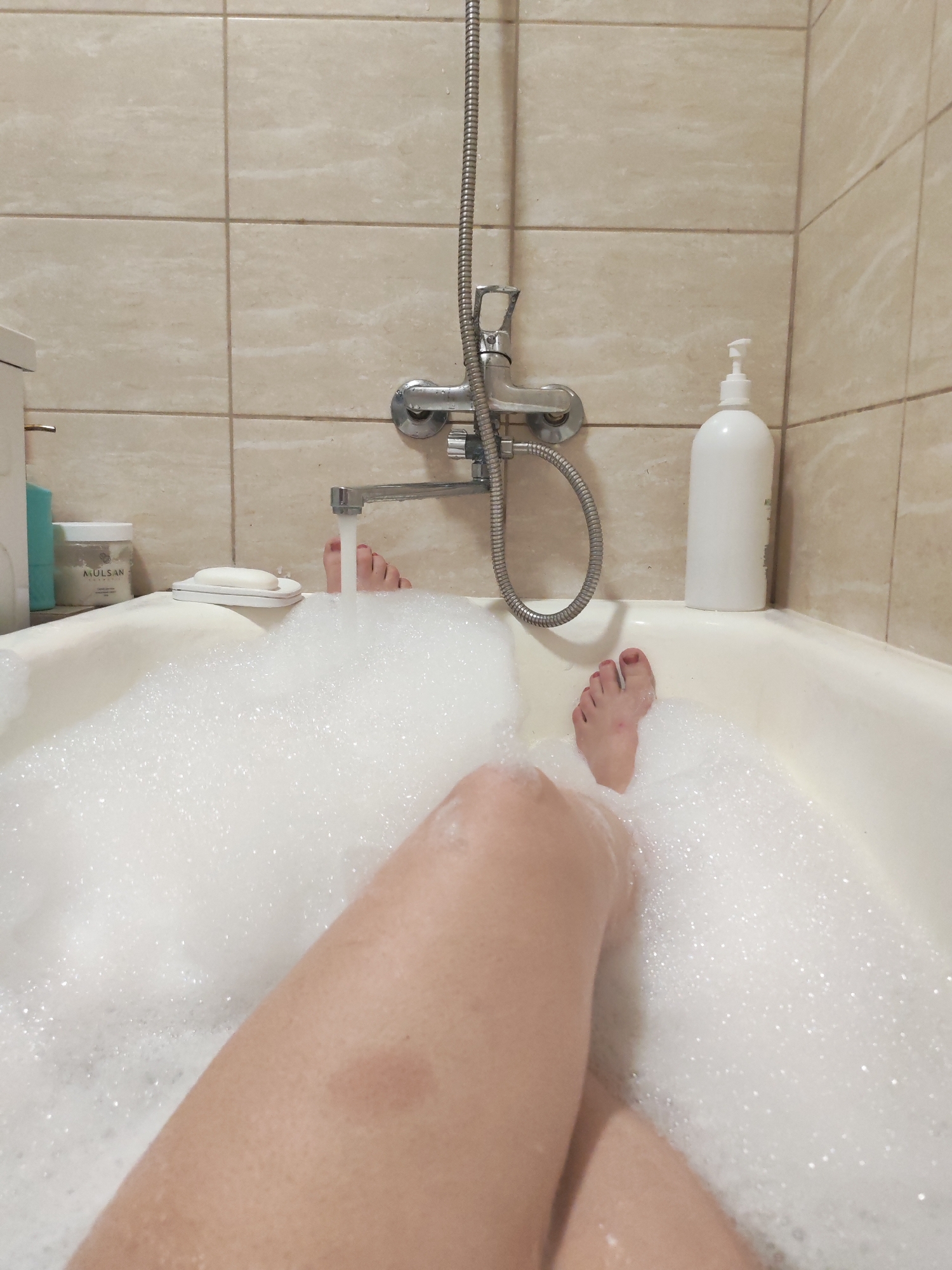 The image shows a person in a bathtub filled with bubbles. The person's legs are visible in the foreground. Two feet are visible in the middle of the image. Bathroom tiles are visible in the background. A shower faucet, a white bottle of soap, and other toiletries are positioned near the edge of the tub. The photo does not provide enough information to determine the location is in Izhevsk, Russia.