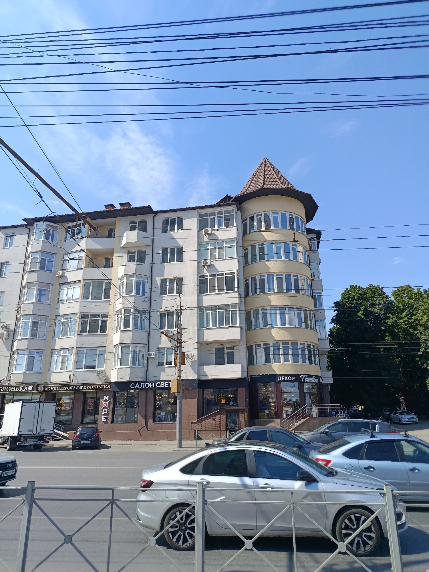 This is an eye-level, medium shot captured on a clear, sunny day, showcasing a multi-story building on a city street in Saratov, Russia. The building features a distinctive round tower with a conical roof, juxtaposed with a more conventional rectangular facade. The architecture is characterized by numerous windows and balconies, with air conditioning units visible on the exterior. A truck is parked on the street, and several cars are driving or parked nearby. Overhead, a network of electrical wires crosses the sky, suggesting an urban environment. The ground level of the building houses several shops, with visible signage in Russian, including "САЛОН СВЕТ" (Salon Svet), "ДЕКОР" (Decor), and "КОНДИТЕРСКАЯ КУЛИНАРИЯ" (Confectionery Culinary). A silver sedan is prominently positioned in the foreground, its rear end facing the camera. A metal railing with a diamond pattern runs horizontally across the lower portion of the image. The overall impression is of a typical street scene in a Russian city.