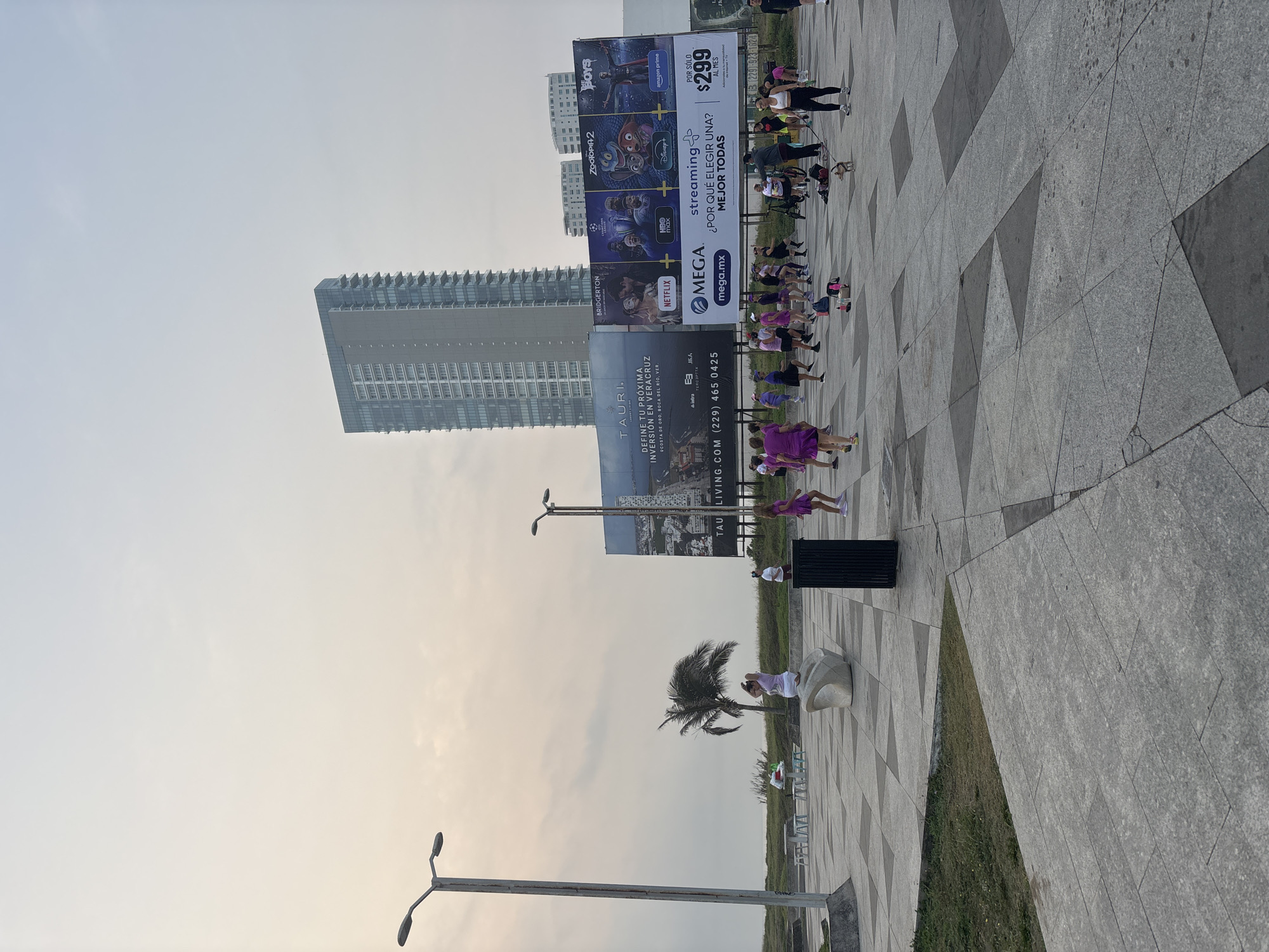 This media file captures an outdoor fitness class or group exercise event taking place on a patterned tiled plaza in Veracruz, Mexico, likely during late afternoon or early evening under a hazy sky.
In the foreground, a group of approximately fifteen to twenty people, mostly women dressed in activewear (some in purple attire), are engaged in a synchronized exercise activity, resembling a dance or aerobics class. They are spread across the patterned grey and dark grey tiled ground. A black rubbish bin is visible near them. Further back, a solitary palm tree stands beside a modern, sculptural white bench.
The background features a modern urban landscape with several tall, multi-story buildings. Dominating the middle ground are two large billboards. The left billboard advertises real estate: "TAURI," "DEFINE TU PRÓXIMA INVERSIÓN EN VERACRUZ," and "COSTA DE ORO, BOCA DEL RÍO, VER.," along with "TAU LIVING.COM (229) 465 0425." The right billboard promotes a streaming service called "MEGA," offering various platforms like Netflix (with "BRIDGERTON"), HBO Max (with a Champions League logo), Amazon Prime (with "BOYS"), Disney+ (with "ZOOTOPIA 2"), and asking "¿POR QUÉ ELEGIR UNA? MEJOR TODAS" (WHY CHOOSE ONE? ALL THE BEST), for "POR SÓLO $299 AL MES" (FOR ONLY $299 PER MONTH). Two streetlights are also visible, one in the lower left foreground and another further back on the left side, adding to the urban setting. Sections of green grass line the edges of the tiled plaza.