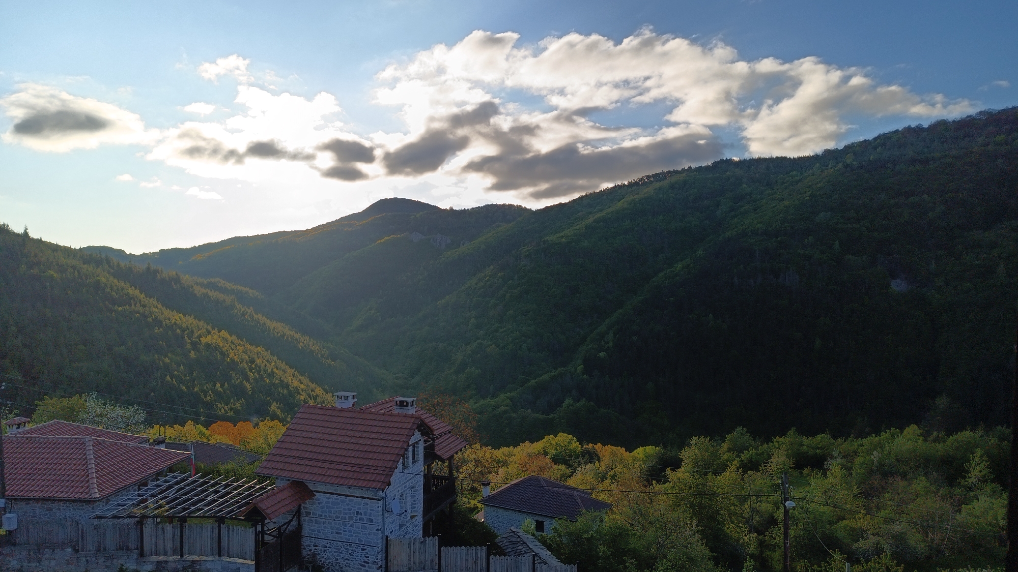 The image shows a landscape in the city of Vrata, Bulgaria, featuring buildings in the foreground and mountains in the background under a sky with clouds. The buildings have red tile roofs and stone walls. There are trees with green and yellow leaves scattered around and behind the buildings. The mountains are covered in green forest. The sky is blue with clouds, and the sun is partially obscured by the clouds. The lighting suggests it is either early morning or late afternoon.