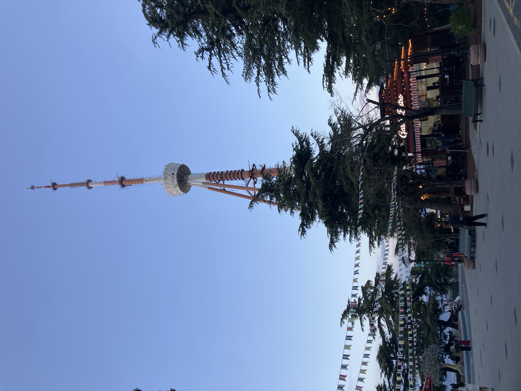 The image captures an outdoor scene in Tbilisi, Georgia, centered around the distinctive Tbilisi TV Tower. The tall, white and reddish-brown tower dominates the left side of the frame, featuring a multi-level circular observation deck or structure near its top, and segmented sections along its shaft. The sky above is clear and bright blue, suggesting good weather.
The setting appears to be a park or promenade, characterized by abundant dark green coniferous trees that frame the top and right portions of the image, casting some shade. On the right, nestled among the trees, is a cozy-looking cafe or bistro with a red and white striped awning and numerous warm string lights illuminating its facade. Visible text on the cafe sign reads "Gora Bistro".
Strings of colorful, rectangular bunting flags are strung horizontally across the scene, adding a festive touch. A paved pathway with light-colored square patterns extends into the distance on the right, where a few blurry figures of people can be seen walking or standing. One person is clearly visible near a green park bench next to the cafe.
The time of day appears to be late afternoon or early evening, indicated by the clear sky contrasting with the illuminated lights of the bistro, suggesting dusk is approaching. The overall atmosphere is pleasant and inviting, depicting a popular public space.