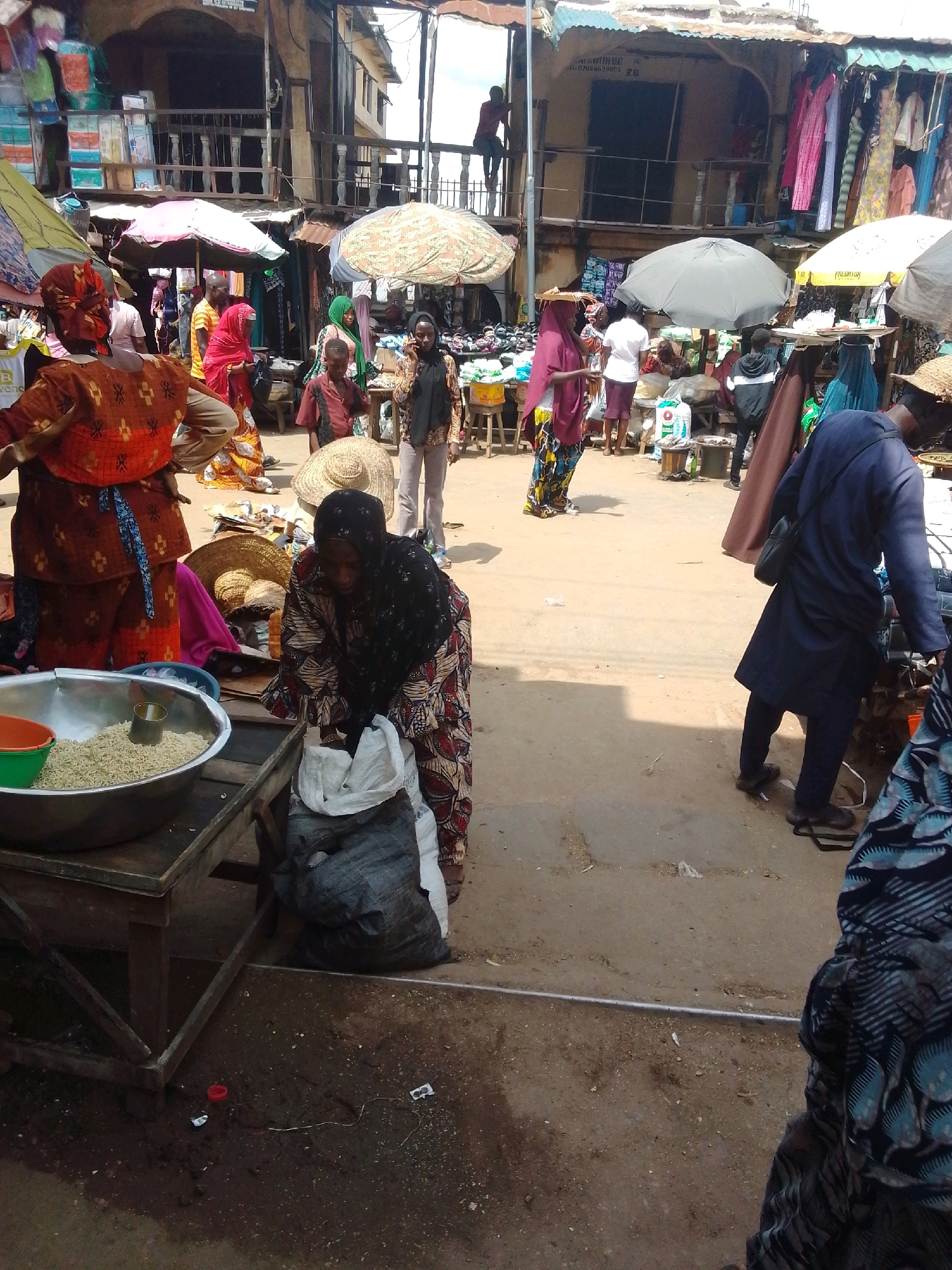 The image depicts a busy street market scene in Okene, Nigeria. In the foreground, a woman in a colorful patterned dress and headscarf is attending to a large bag of goods next to a table. A large metal bowl filled with what appears to be small food items is on the table. To the left, another woman in an orange and brown outfit is partially visible. In the background, various vendors are set up with stalls displaying clothing and other merchandise, shaded by umbrellas and awnings. Several people are seen walking and interacting within the market. The ground is dusty and appears to be a mix of dirt and some paved areas, with scattered debris. The architecture in the background suggests a bustling urban environment.